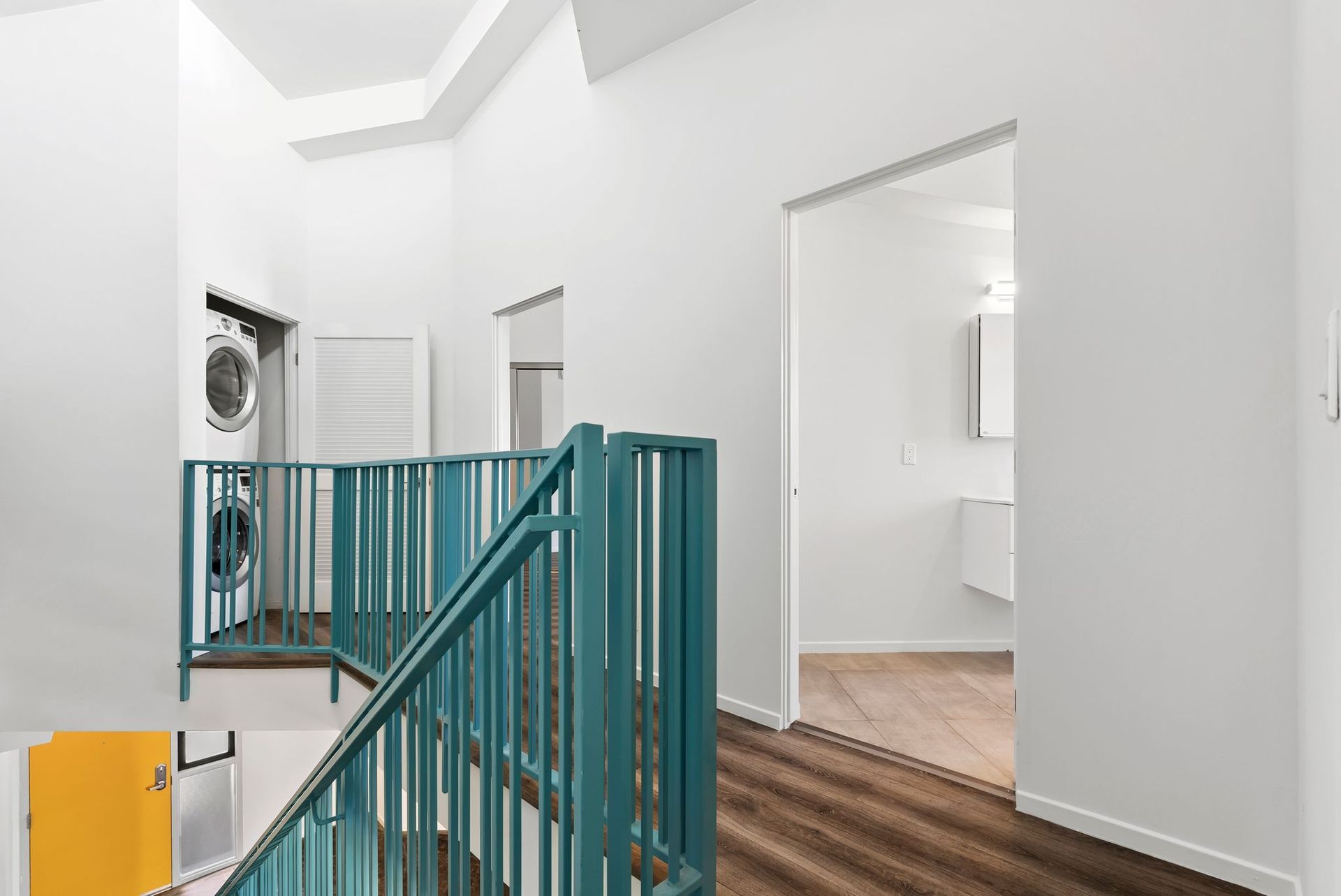 Bright white hallway with teal staircase railing and open doorway to a hardwood room