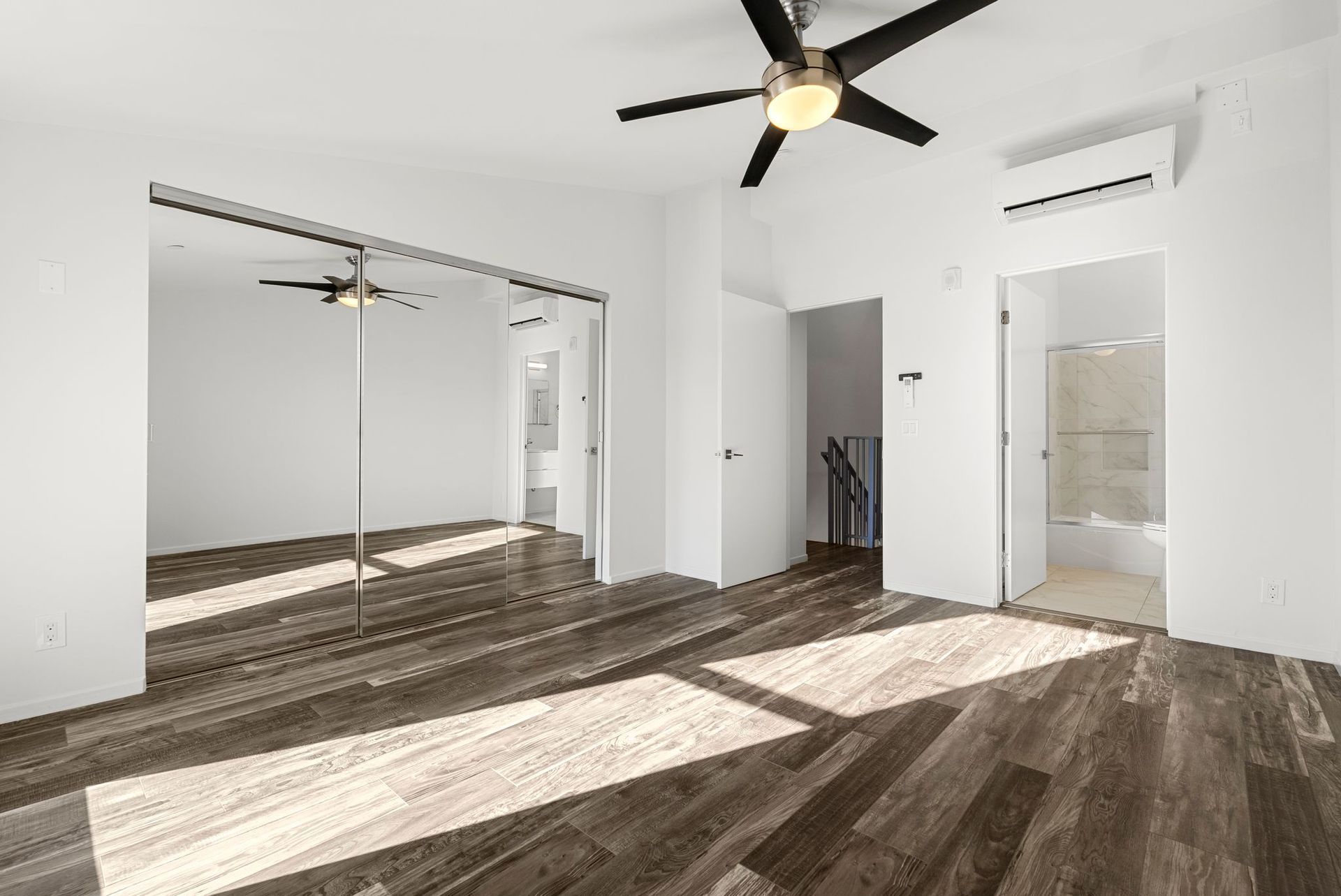 Empty bedroom with mirrored closet doors, wooden floors, and ceiling fan. Light streams in.