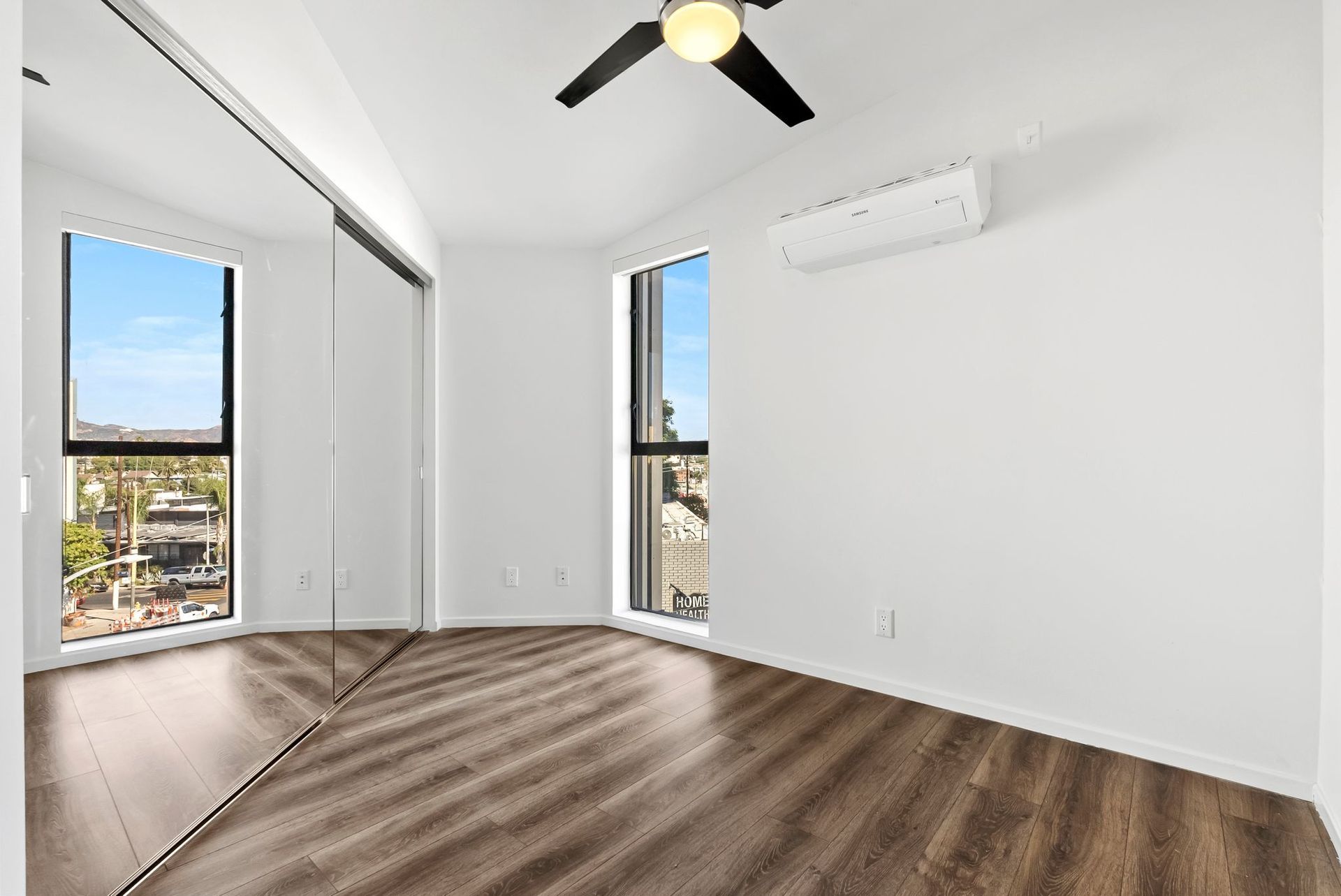 Empty modern bedroom with white walls, wood floors, mirrored closet, ceiling fan, and wall-mounted AC unit.