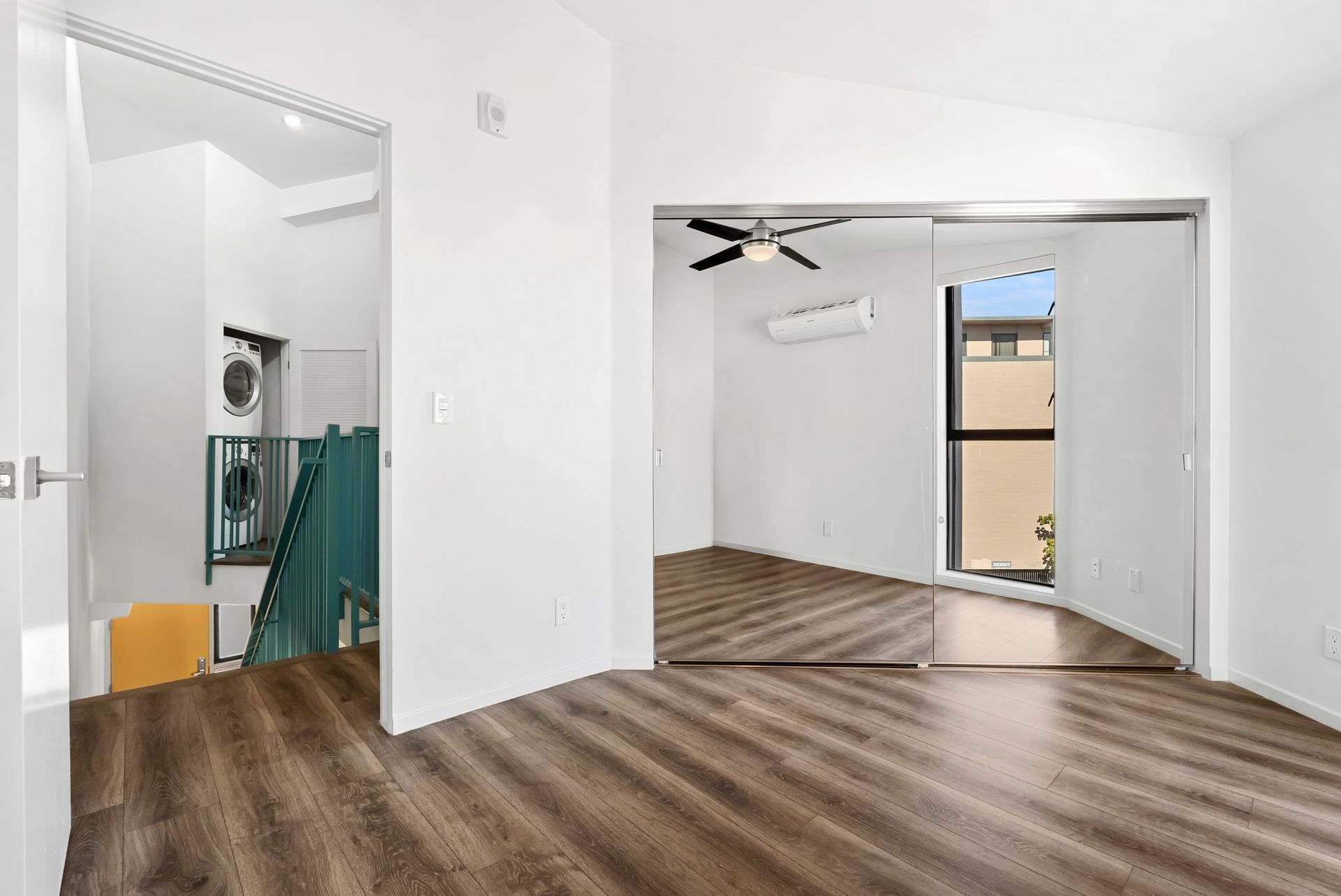 Empty white room with wood floors, ceiling fan, mirrored closet, and open doorway to a bathroom.
