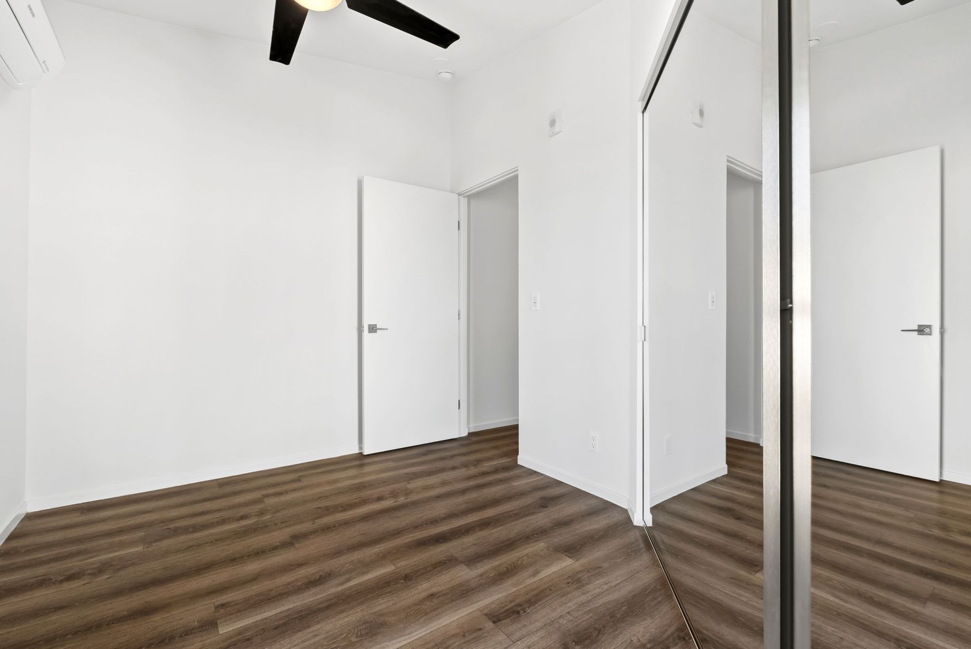 Empty white bedroom with brown carpet, mirrored closet doors, and a ceiling fan