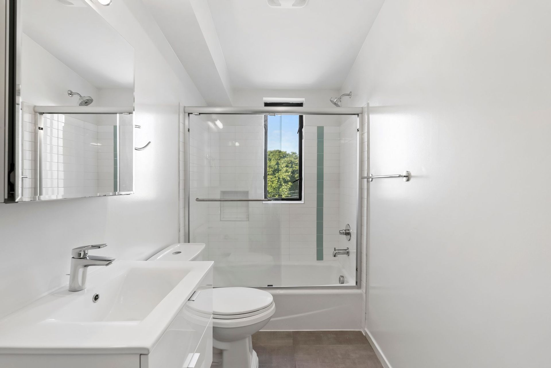 Bright modern bathroom with white vanity, toilet, glass shower, and window view of greenery