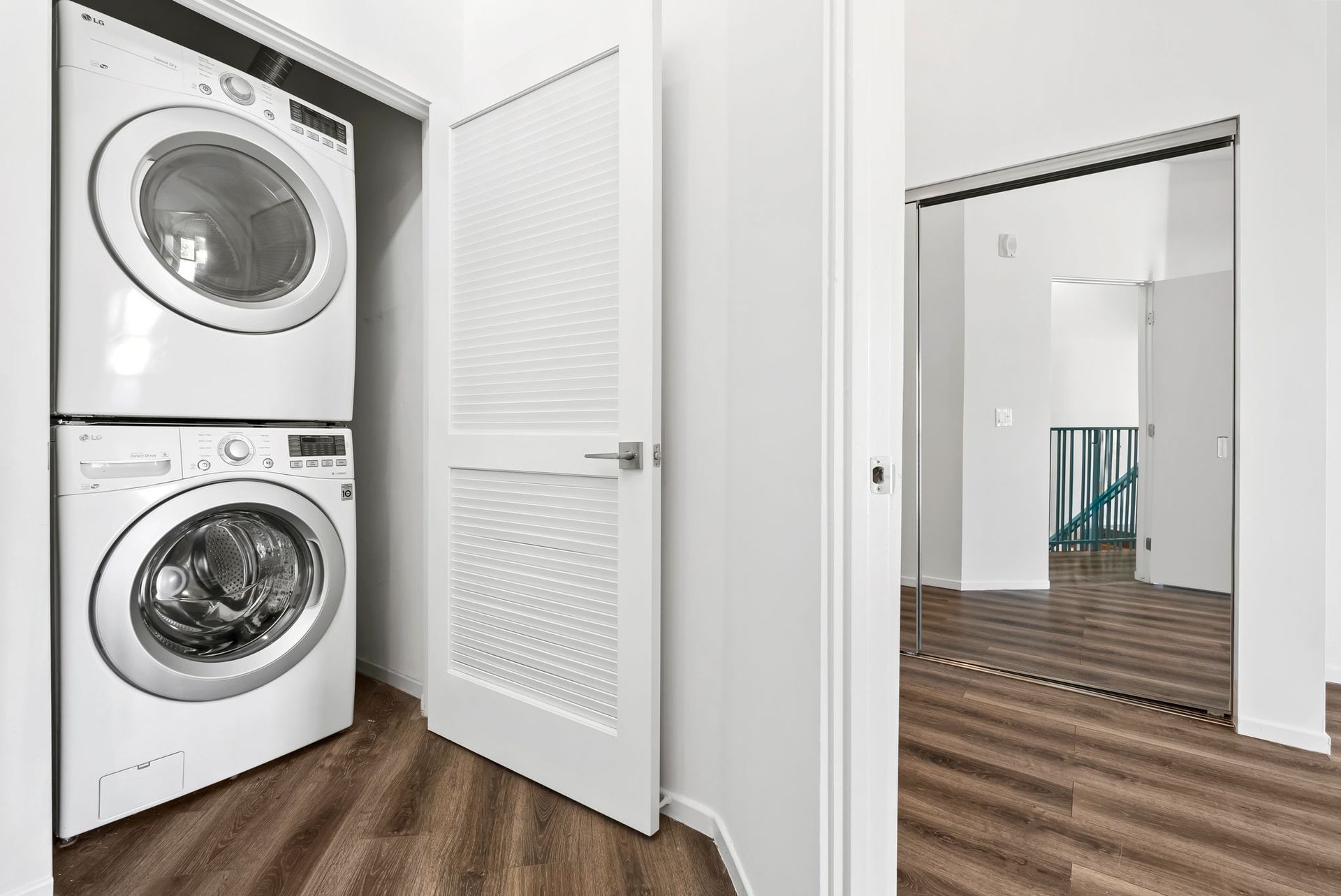 Stacked washer and dryer in a white laundry closet, with wood flooring and an open door nearby