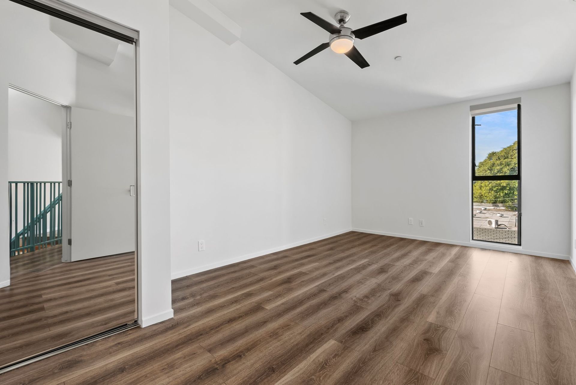 Empty modern room with white walls, wood-look flooring, ceiling fan, and a narrow window overlooking trees
