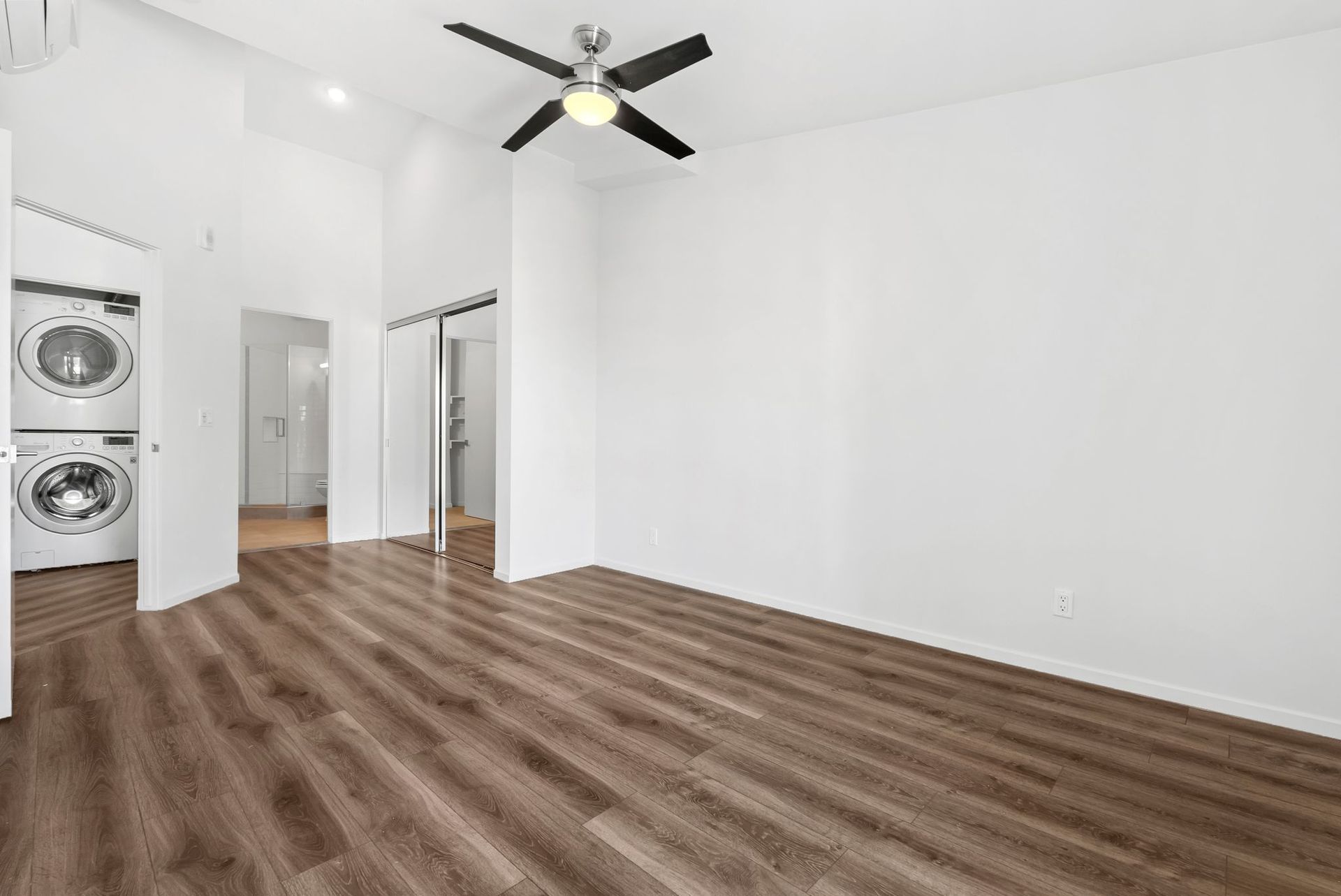 Empty white room with wood flooring, ceiling fan, and stacked washer-dryer in an open closet