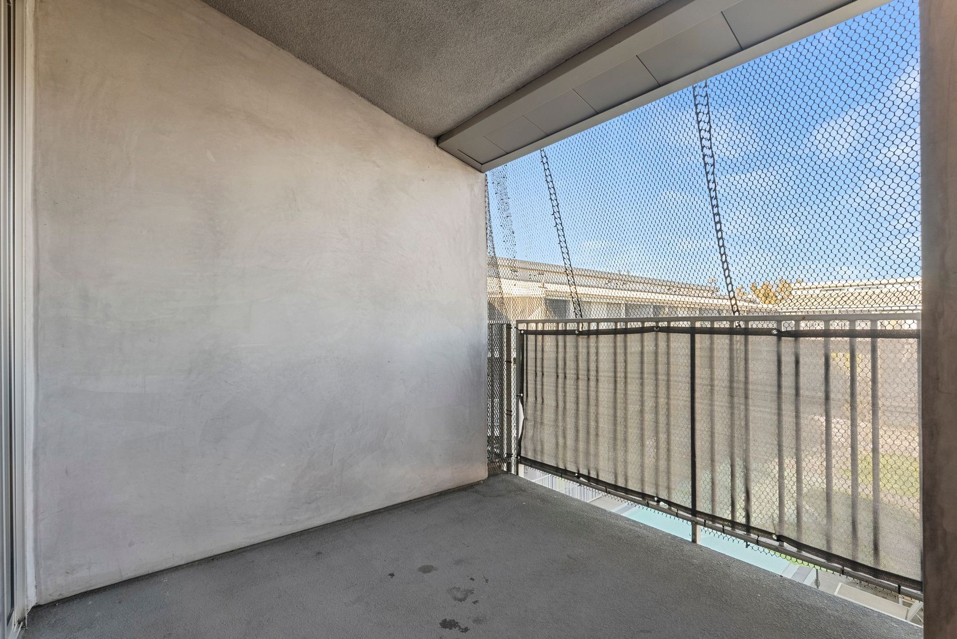 Balcony with gray concrete walls, metal railing, and a mesh screen overlooking a landscape under a blue sky.