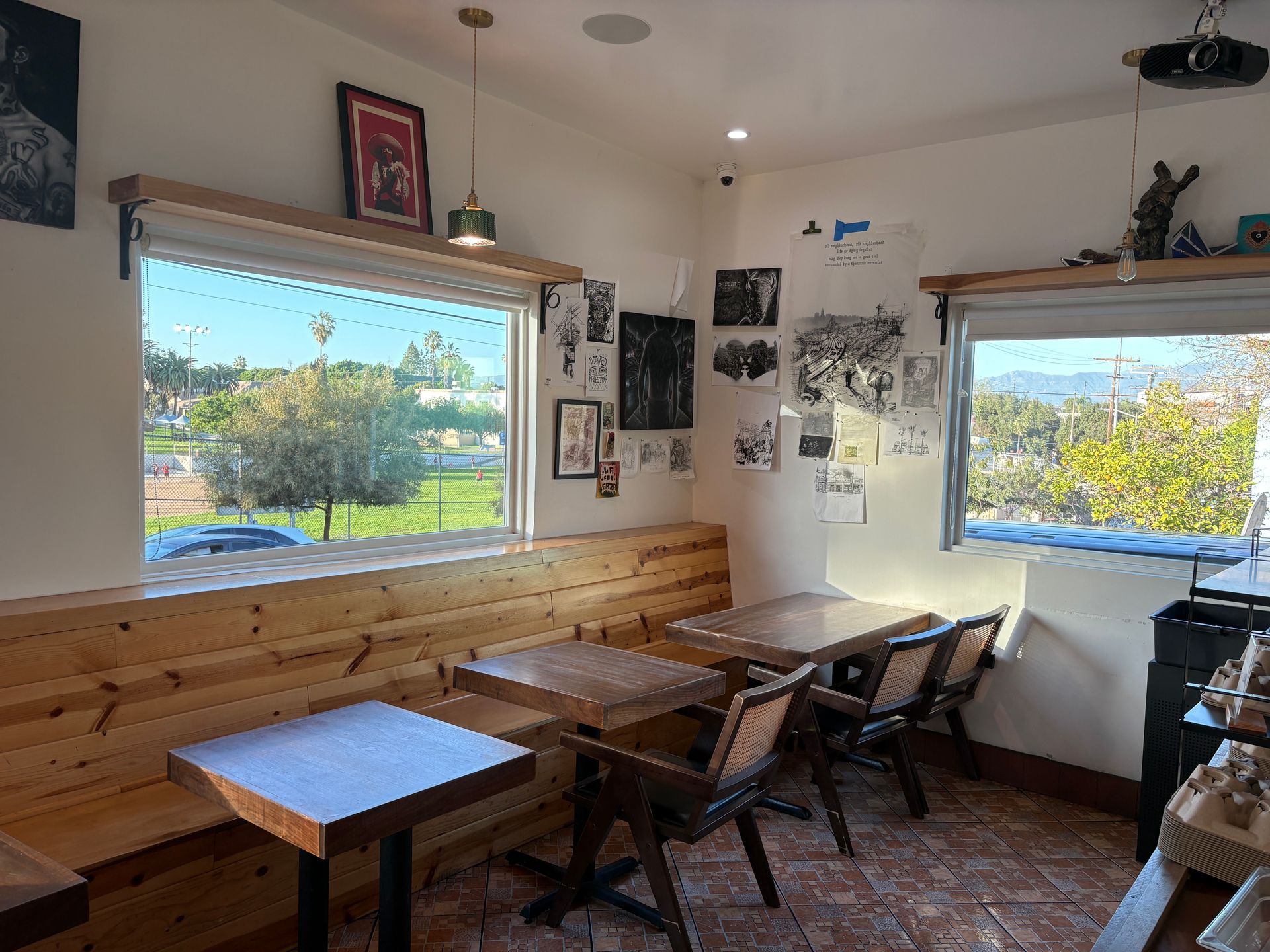 Interior of a cafe with tables, chairs, and windows with outdoor views. Wooden bench seating and artwork on the walls.