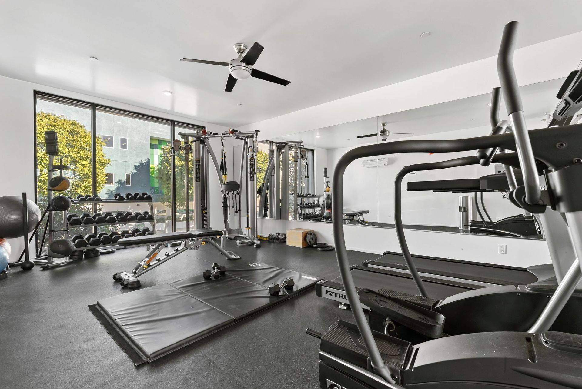Modern home gym with treadmill, cable machine, mats, and large windows.