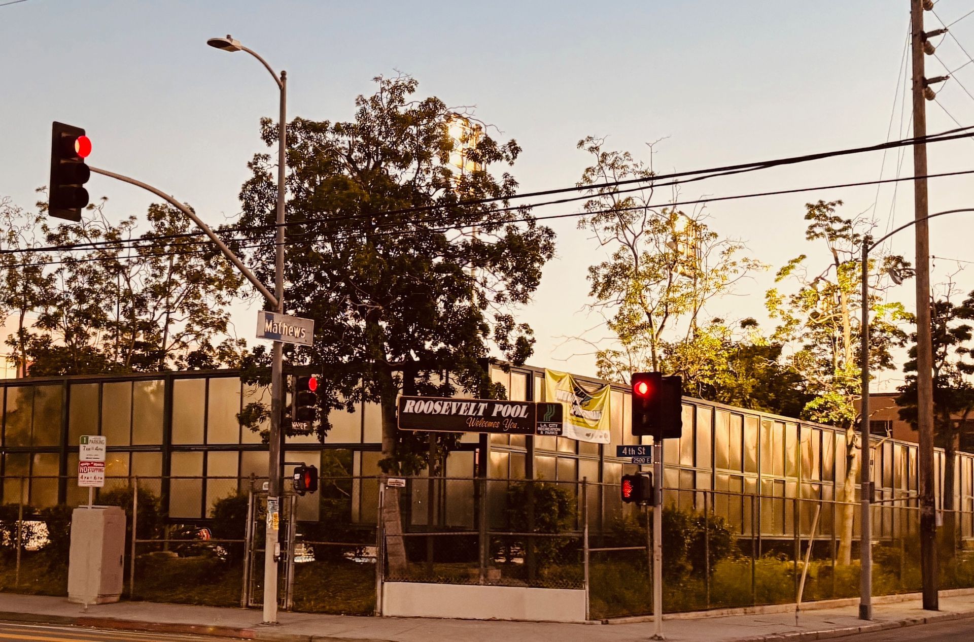 Intersection with traffic lights and a park, framed by a fence and trees, at dusk.