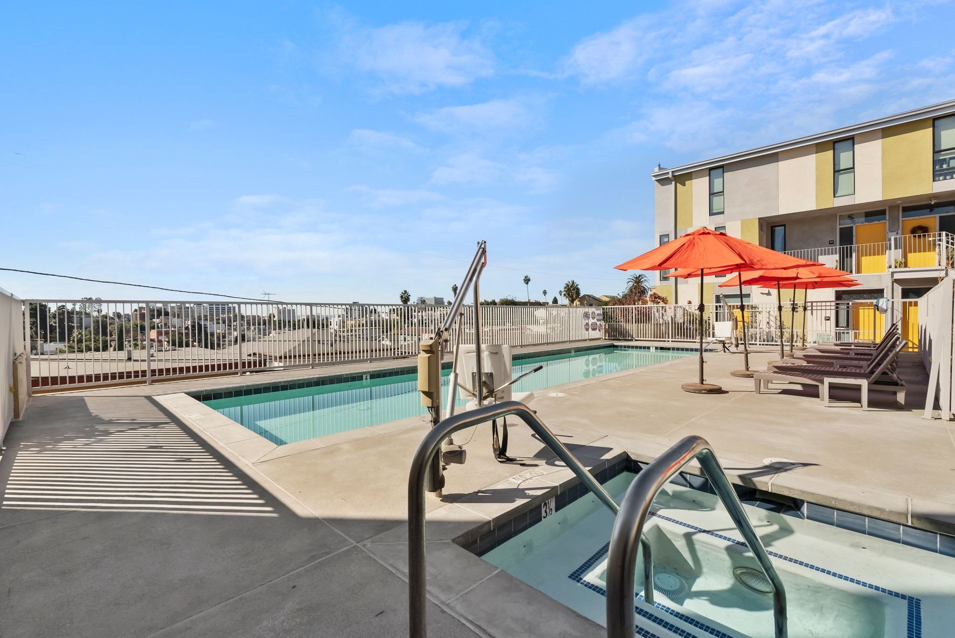 Rooftop pool with lounge chairs, orange umbrellas, and city view under a blue sky