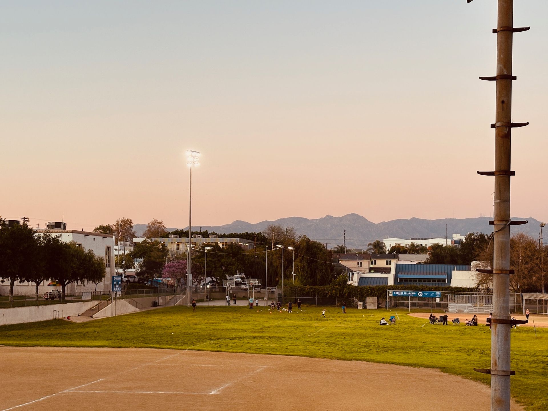 Baseball field at dusk with a lit light tower. Buildings and mountains are visible in the distance.