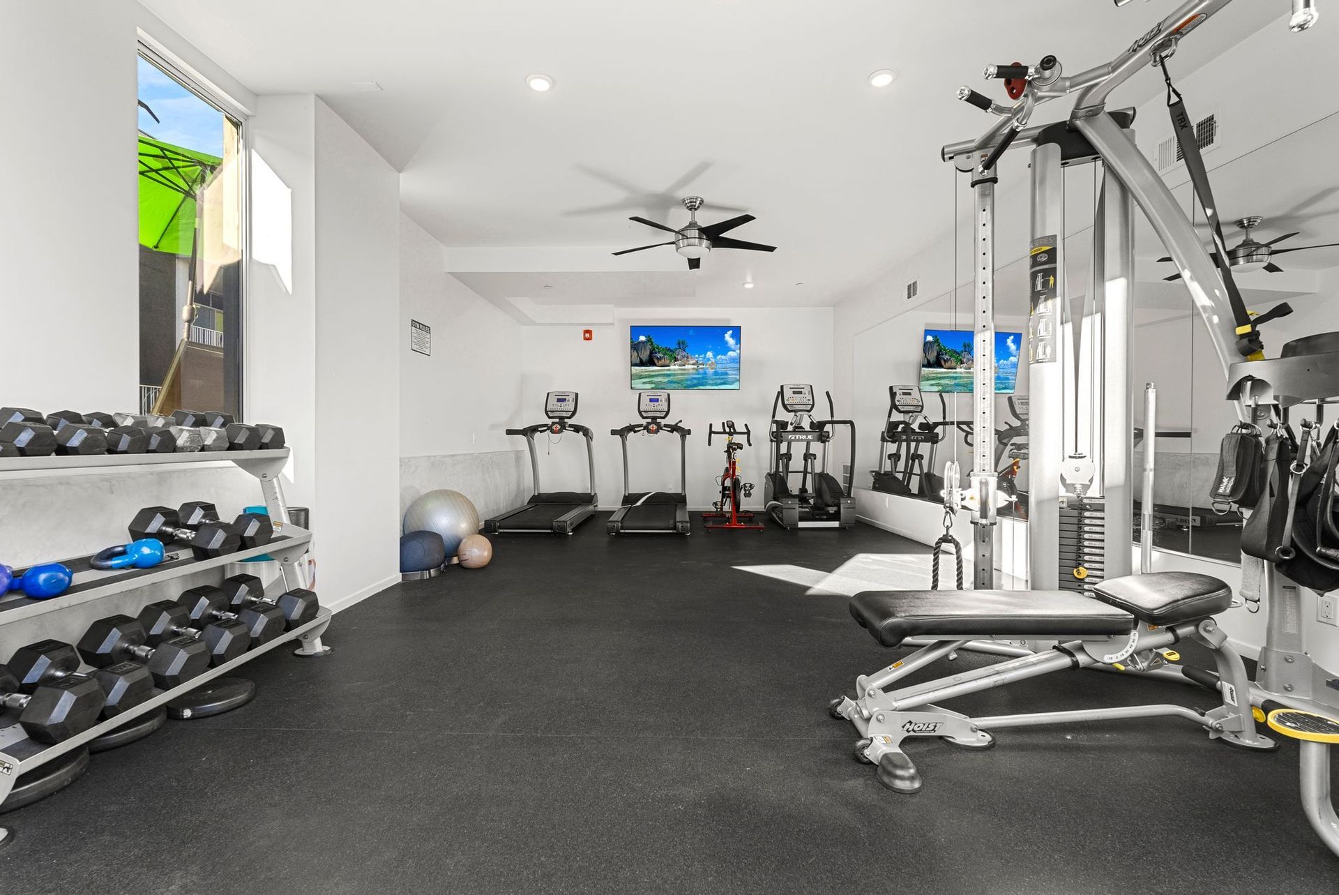 Gym with weight racks, cardio machines, and a multi-station weight machine; neutral-toned, bright.