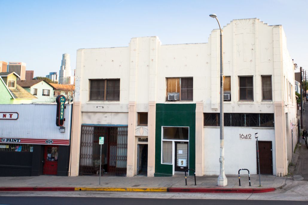 Old storefronts with faded white facades and a green door along a street under a clear sky