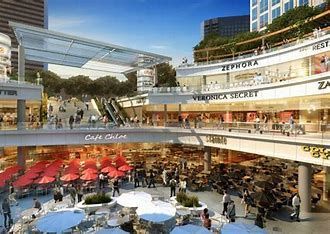 Multi-level outdoor shopping mall with stores, cafes, escalators, and people milling about.
