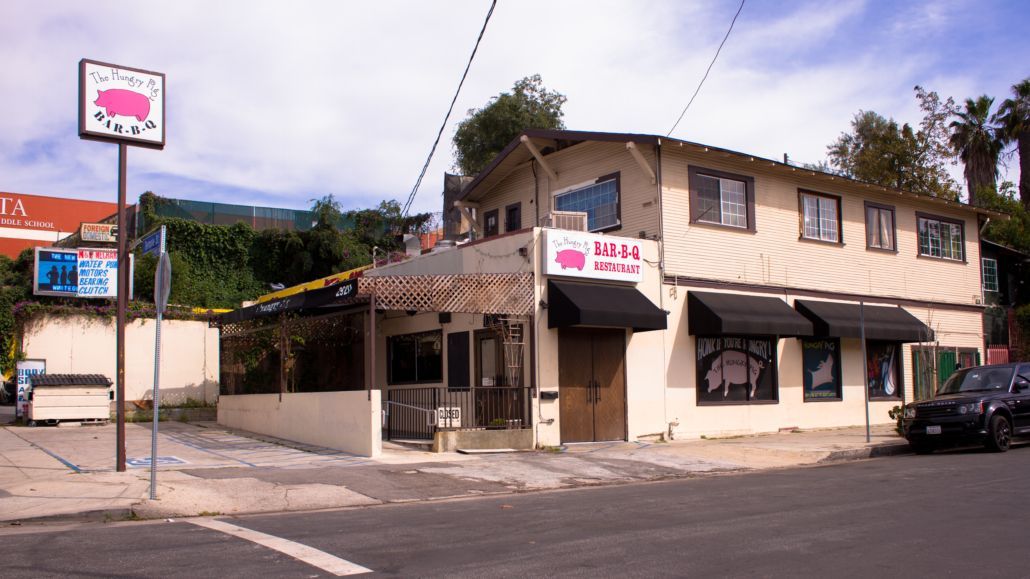 Street corner with a two-story beige building, storefront signs, and a parked car under a partly cloudy sky.