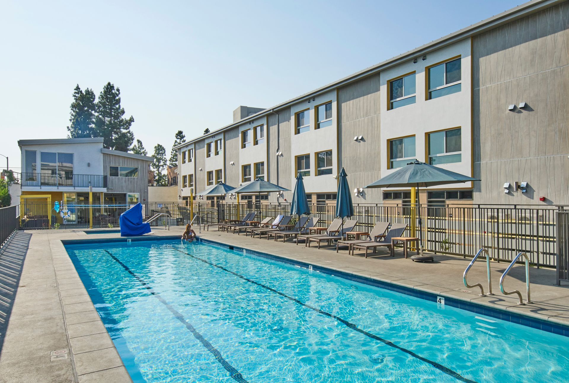 A large swimming pool surrounded by chairs and umbrellas in front of a building.