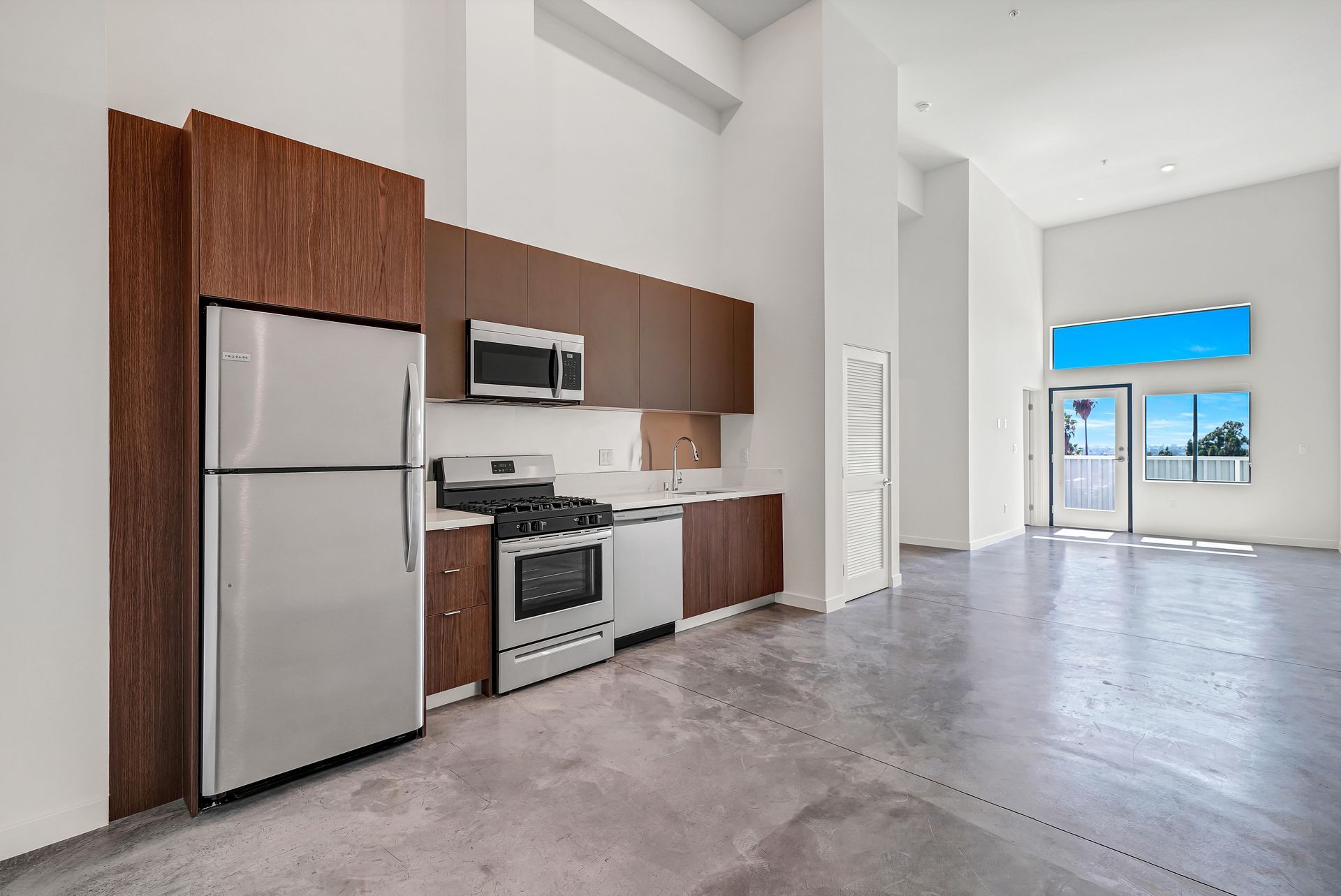 Modern kitchen with stainless steel appliances, dark wood cabinets, and concrete floor. Bright, open space.