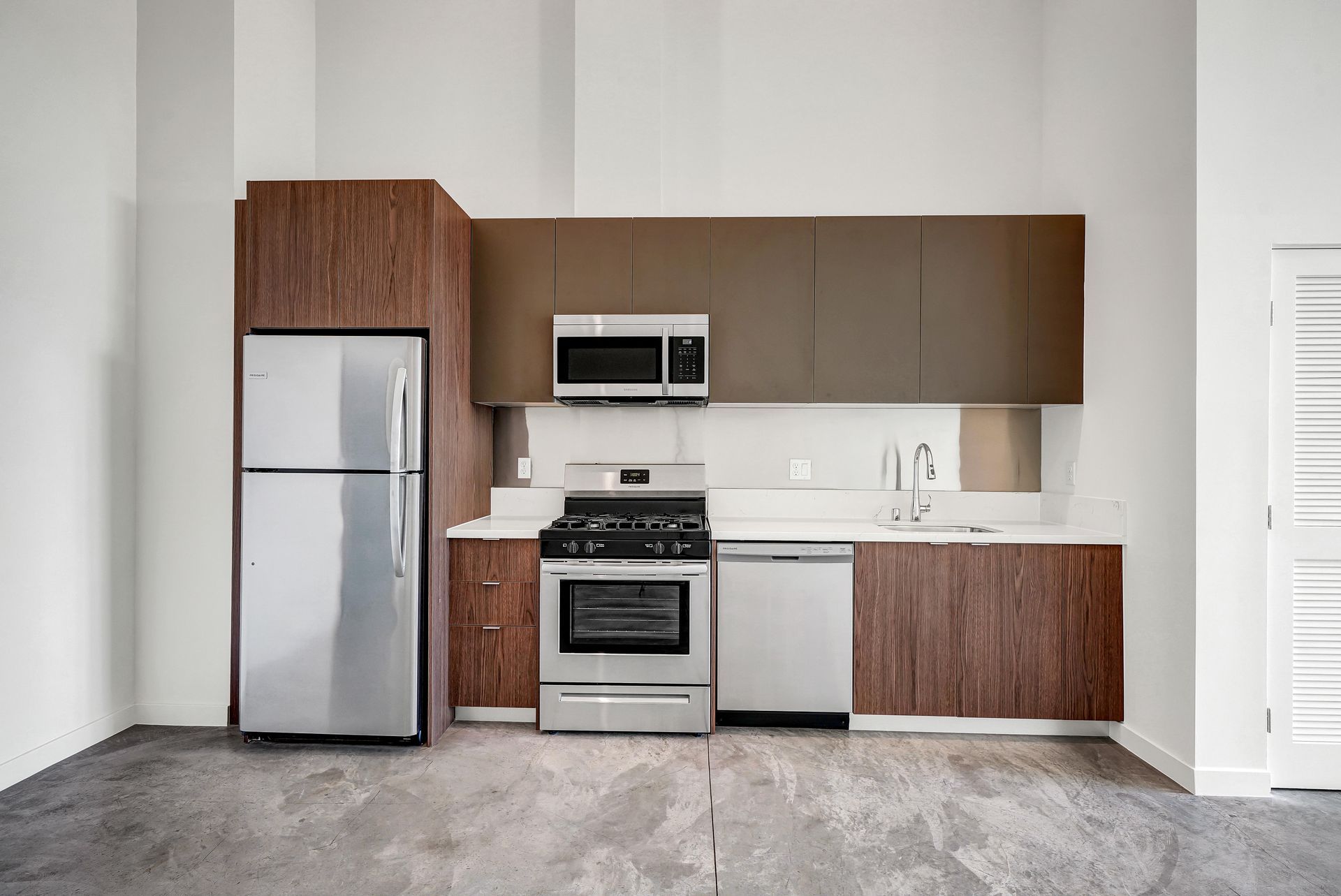 Modern kitchen with stainless steel appliances and brown cabinets. White countertop, concrete floor, and white walls.