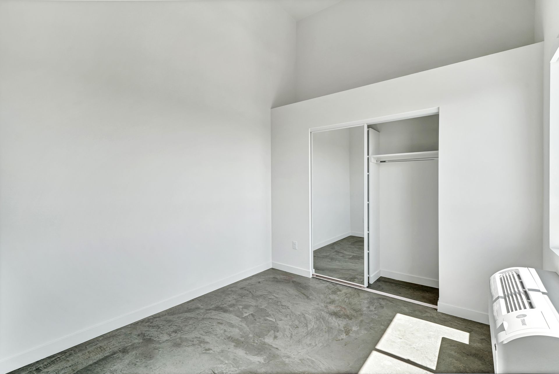 Empty room with concrete floor, white walls, closet with mirrored doors, and a window with sunlight.
