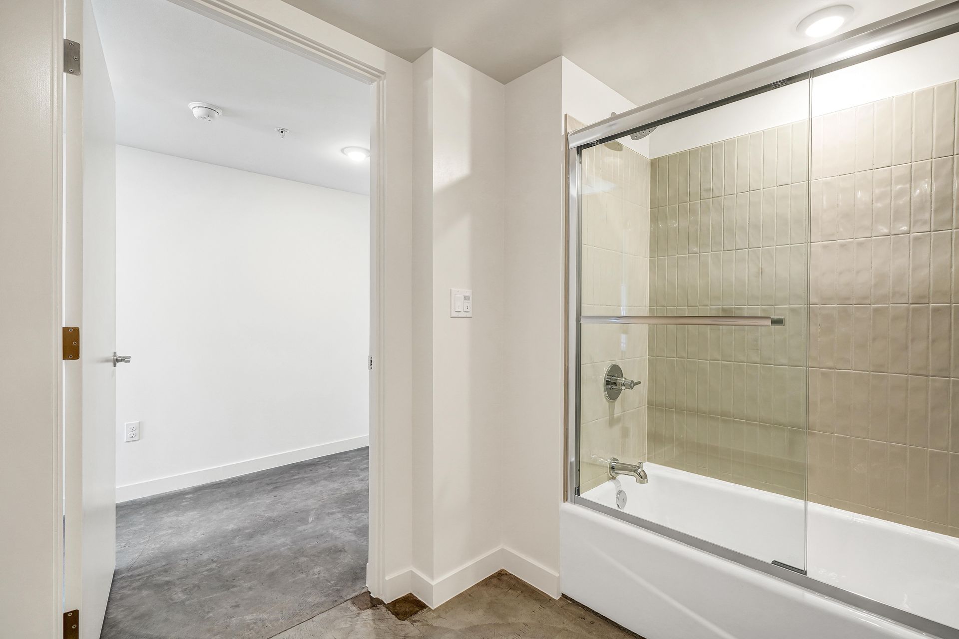 Bathroom with glass shower door, white walls, and a doorway leading to a bedroom with gray carpet.
