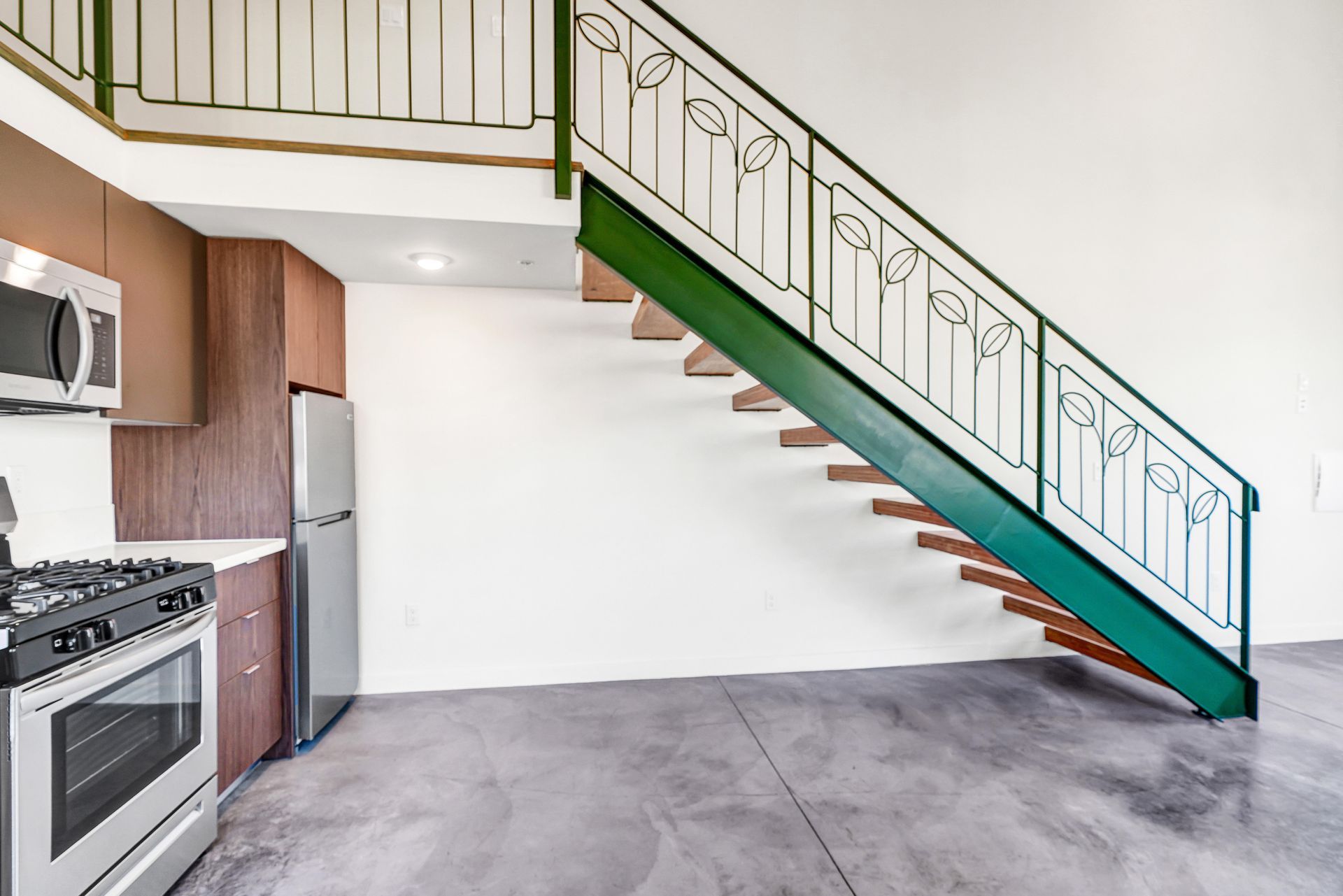 Modern kitchen with stainless steel appliances, stairs leading up, white walls, and concrete floor.