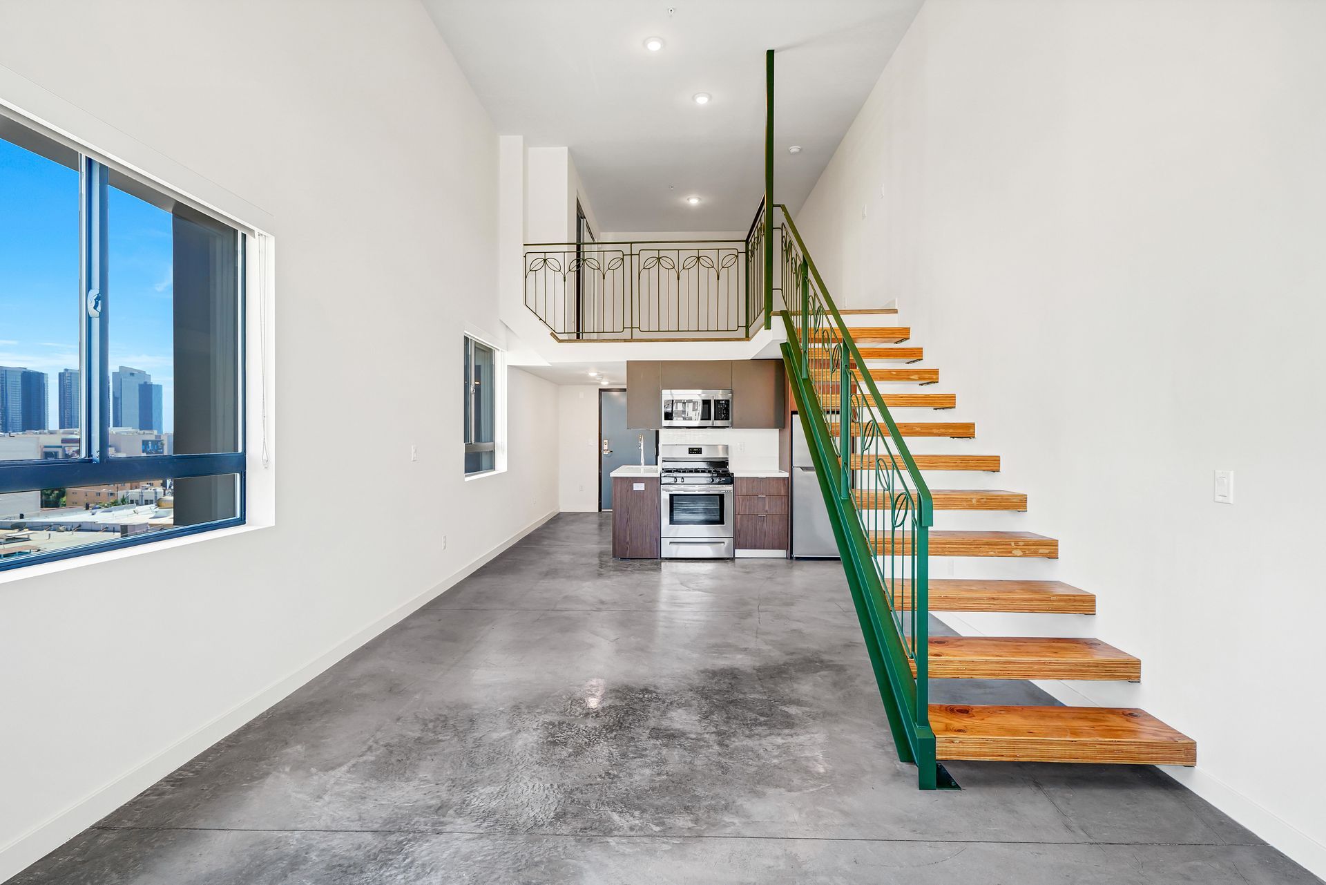 Loft apartment with stairs, kitchen, and mezzanine. Concrete floor, green railing, city view.