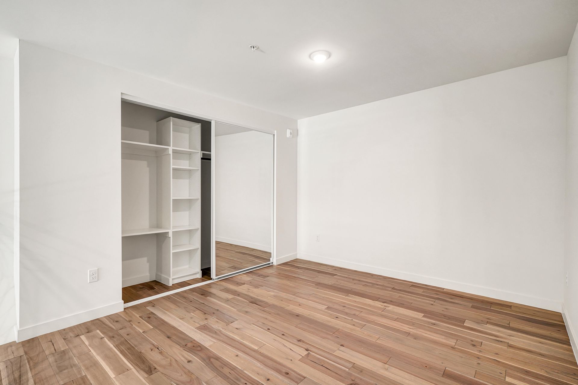 Empty bedroom with hardwood floors, white walls, closet with shelves, and a large mirror.