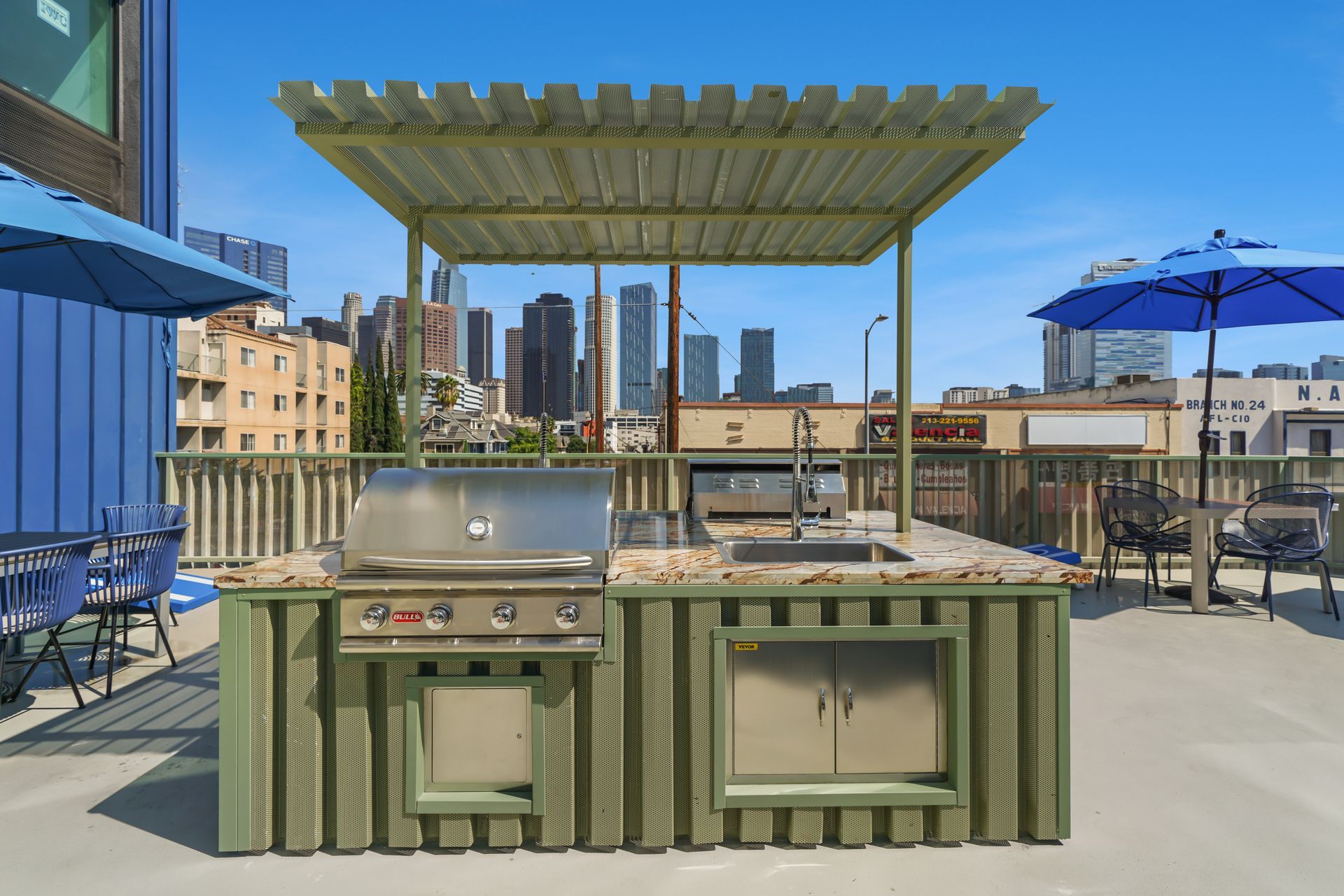 Rooftop outdoor kitchen with grill, sink, cabinets, and awning; cityscape in background.