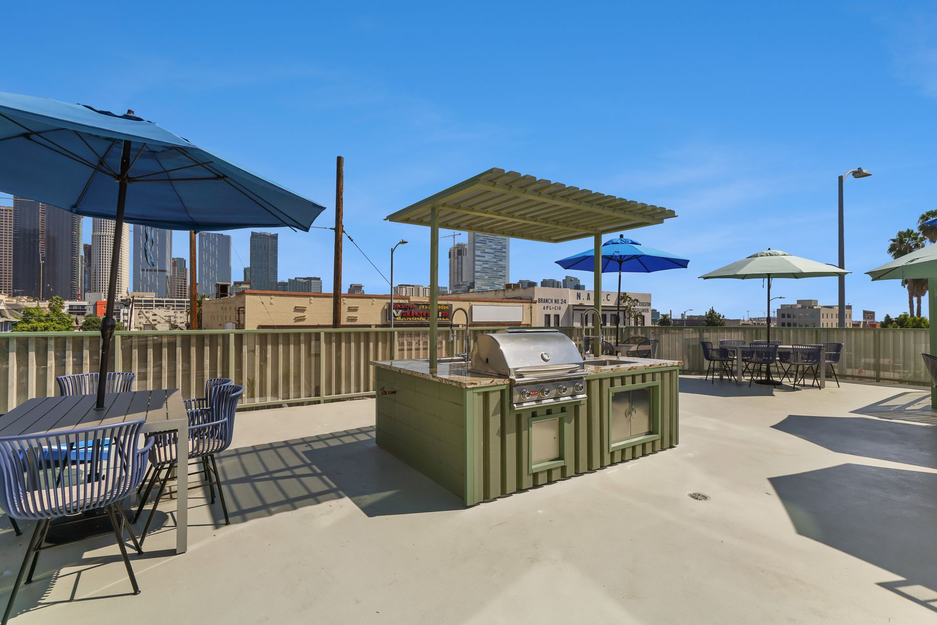 Rooftop patio with grill, tables, and umbrellas. Blue sky and city skyline in the background.