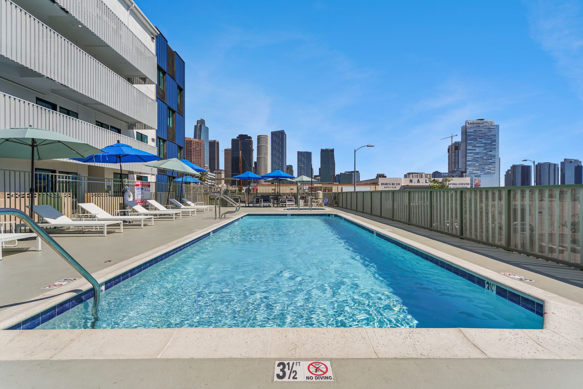 Pool with blue water and umbrellas, a city skyline in the background on a sunny day.