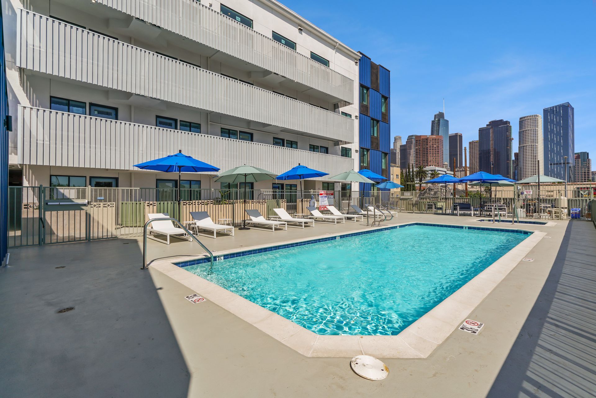 Pool area with blue umbrellas, lounge chairs, and city skyline in the background.