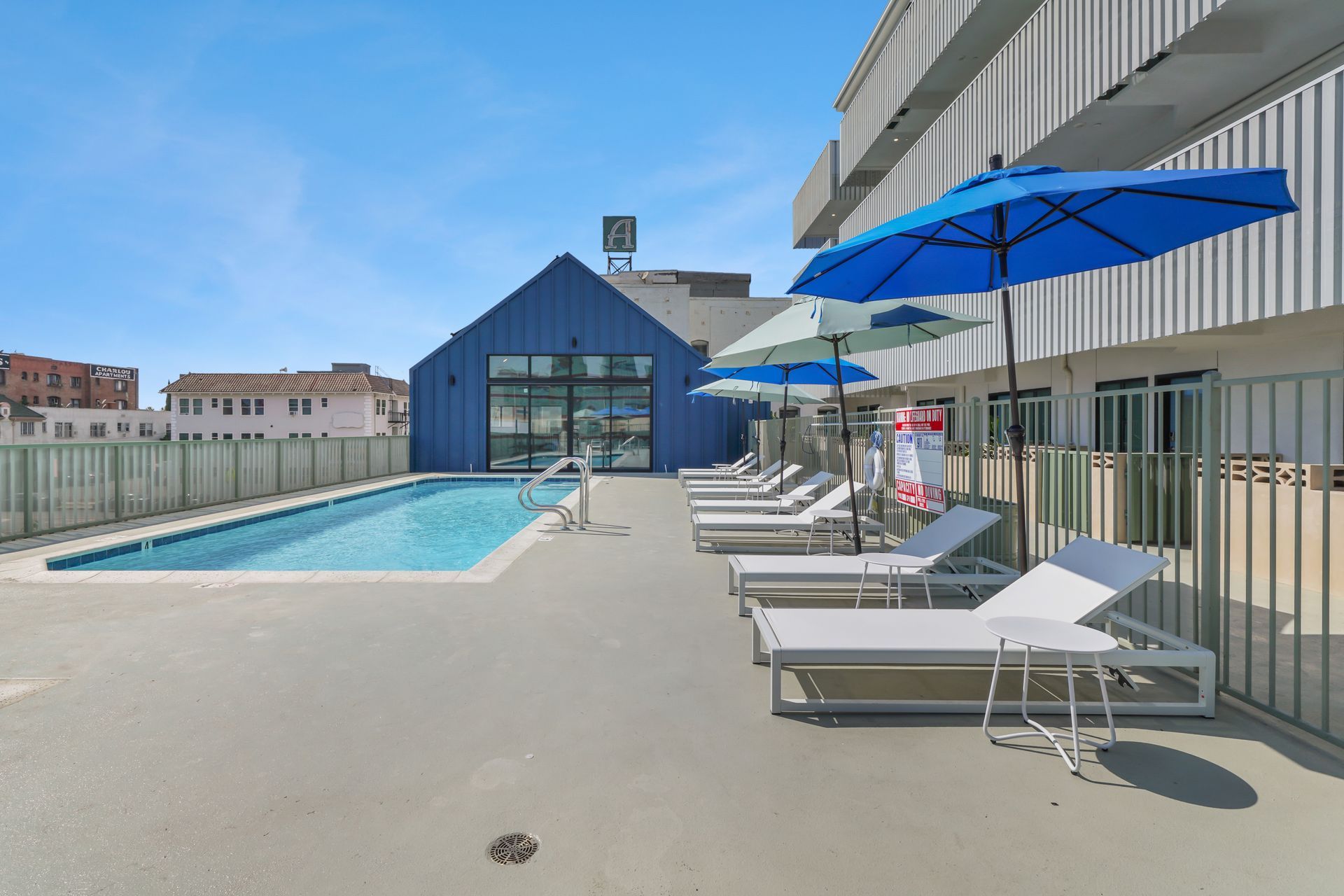 Poolside scene with lounge chairs, blue umbrellas, and a blue building under a bright sky.