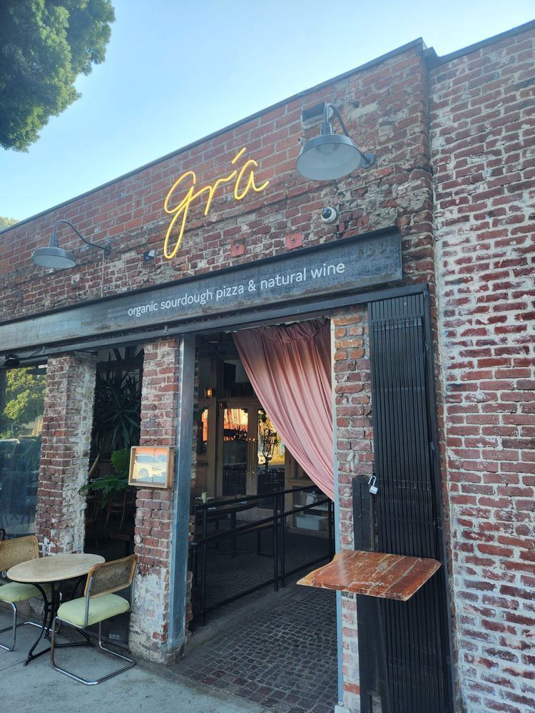 Exterior of Grá restaurant; brick building with neon sign above door, two tables outside.