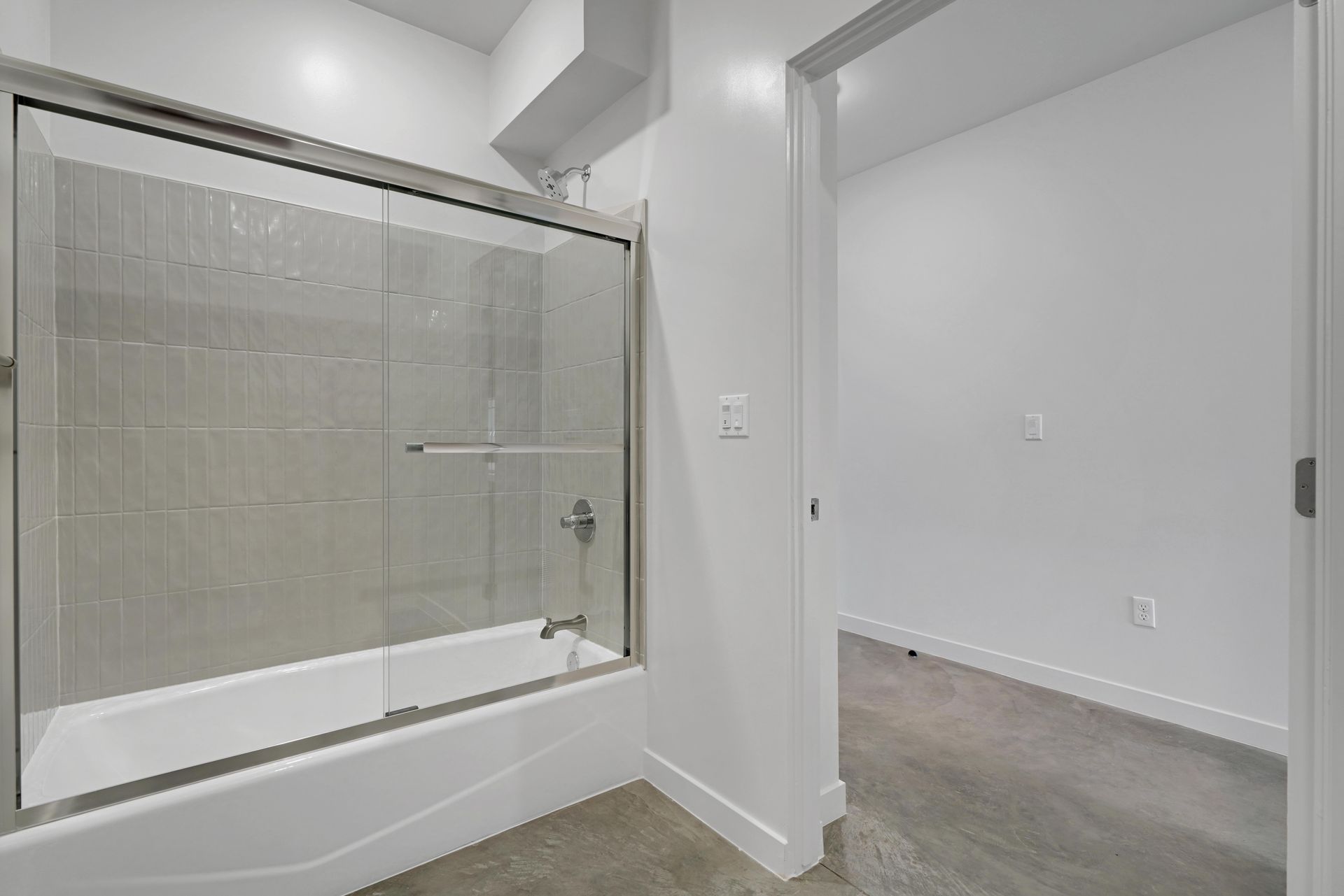 Bathroom with a tub/shower combo, gray tile, glass door, and an adjacent room with a bare floor.