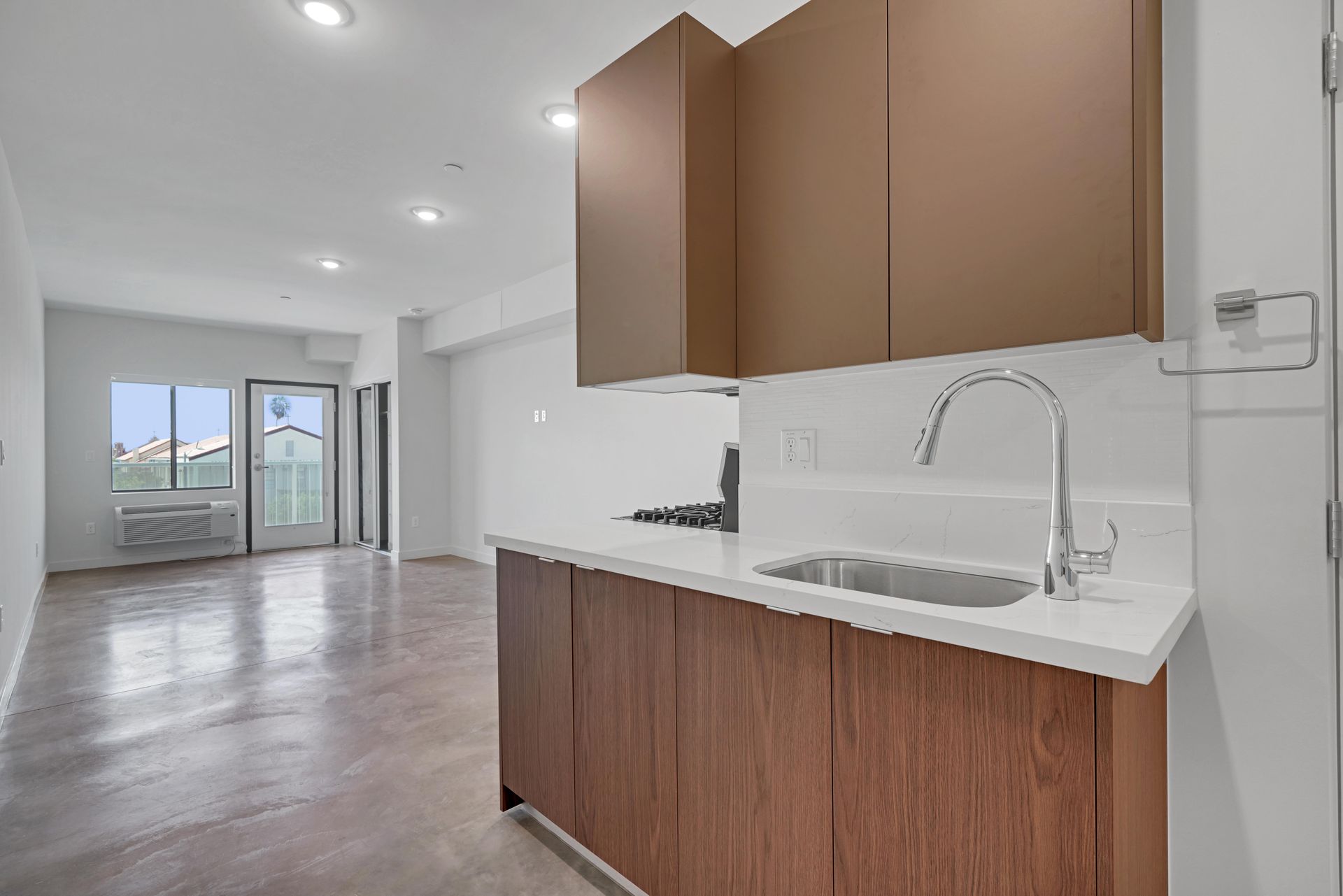 Kitchen area of a modern apartment; brown cabinets, white countertop, sink, and view to a balcony.