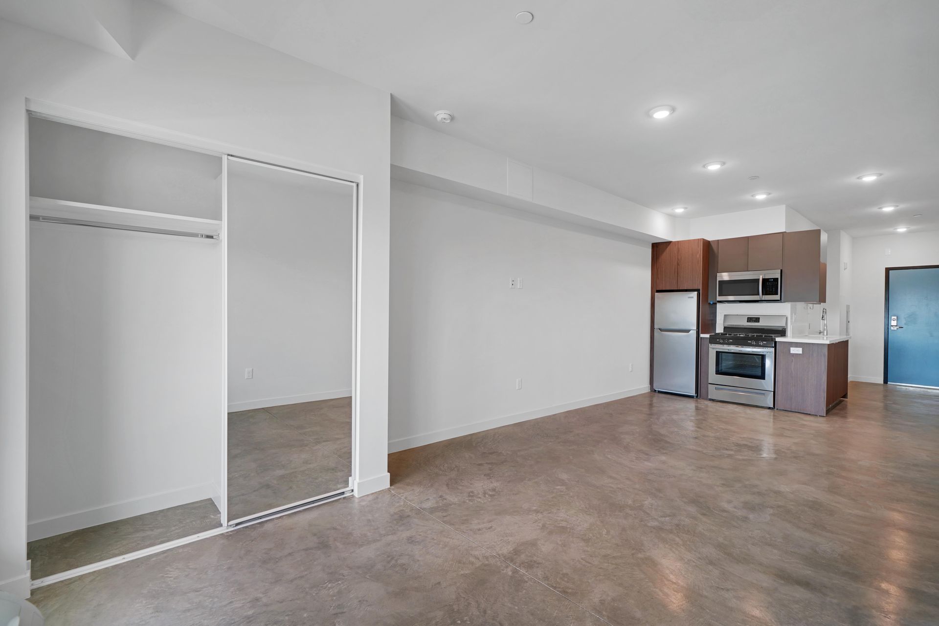 Empty studio apartment with concrete floors, kitchen appliances, and closet.