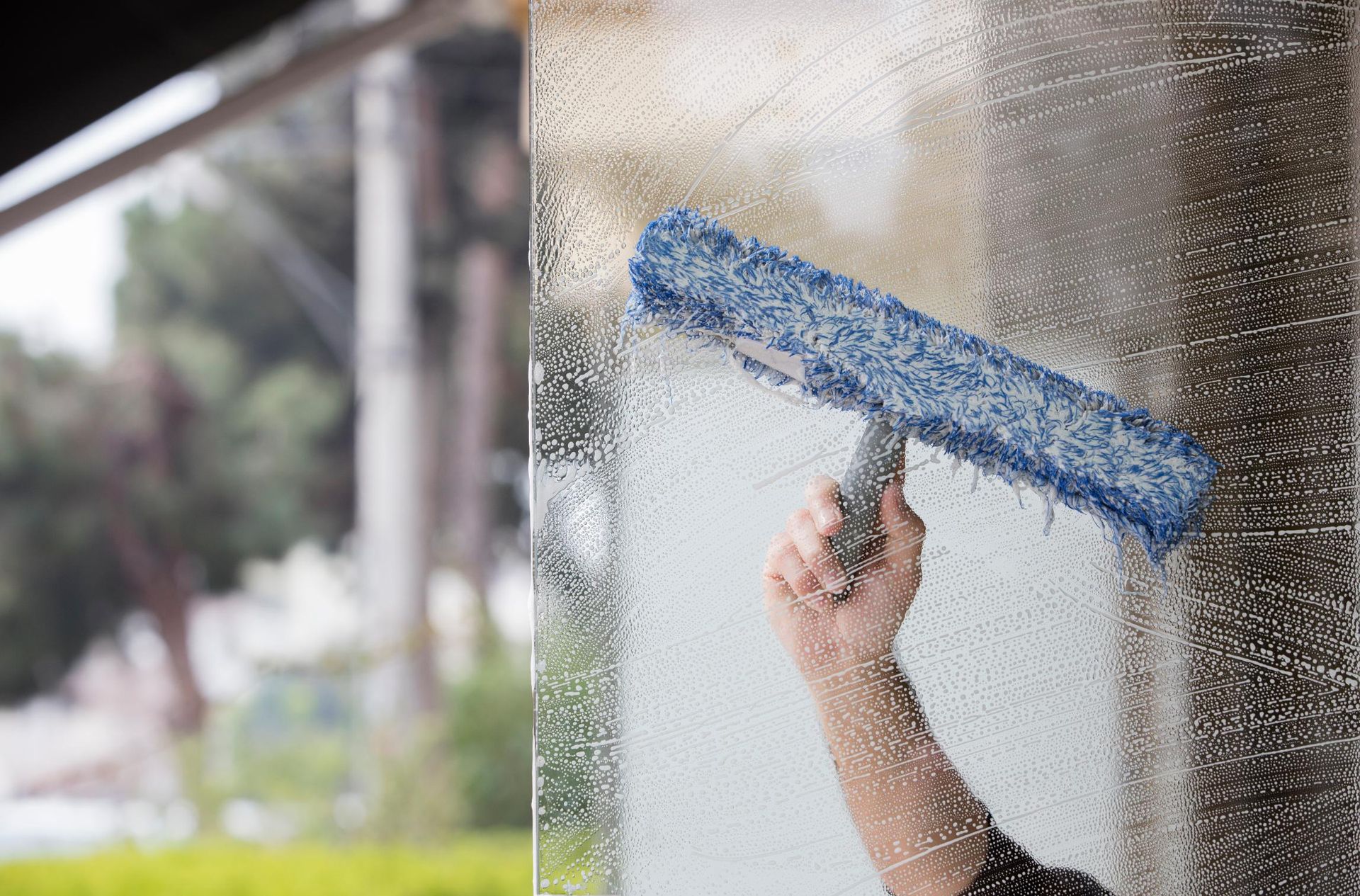 Person using a blue and silver squeegee to clean a wet window. Blurred outdoor background.
