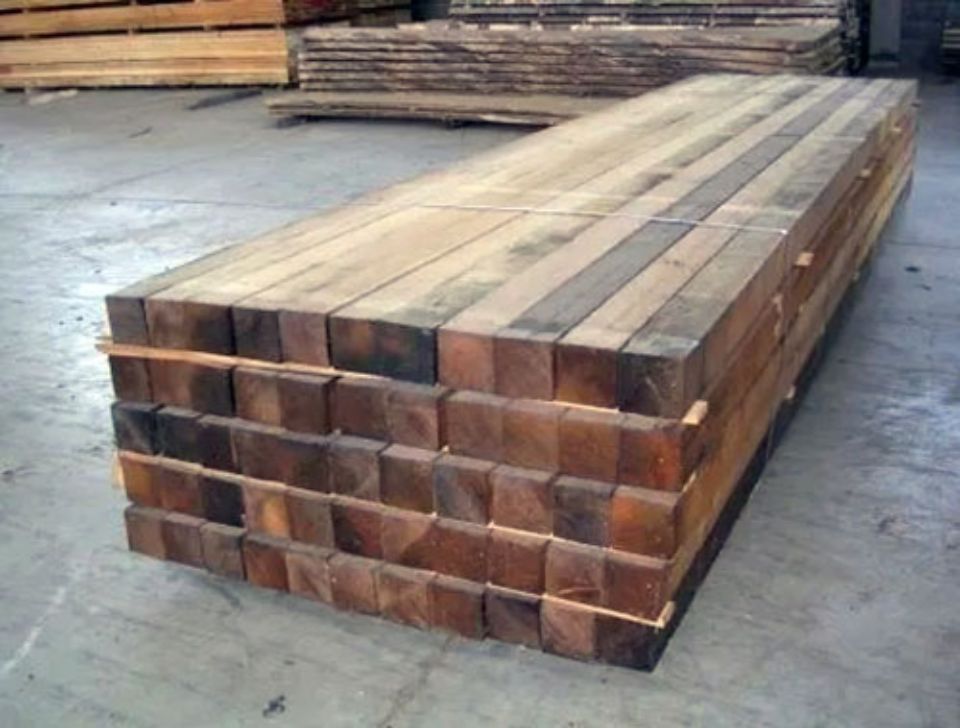 Una pila di travi di legno squadrate, di diverse tonalità di marrone, in un ambiente simile a un magazzino.