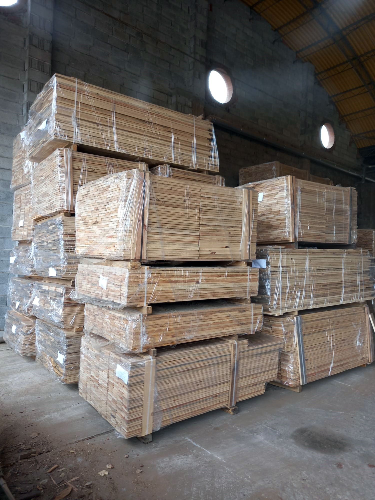Fasci di assi di legno accatastati all'interno di un magazzino, avvolti nella plastica e fissati con delle cinghie.