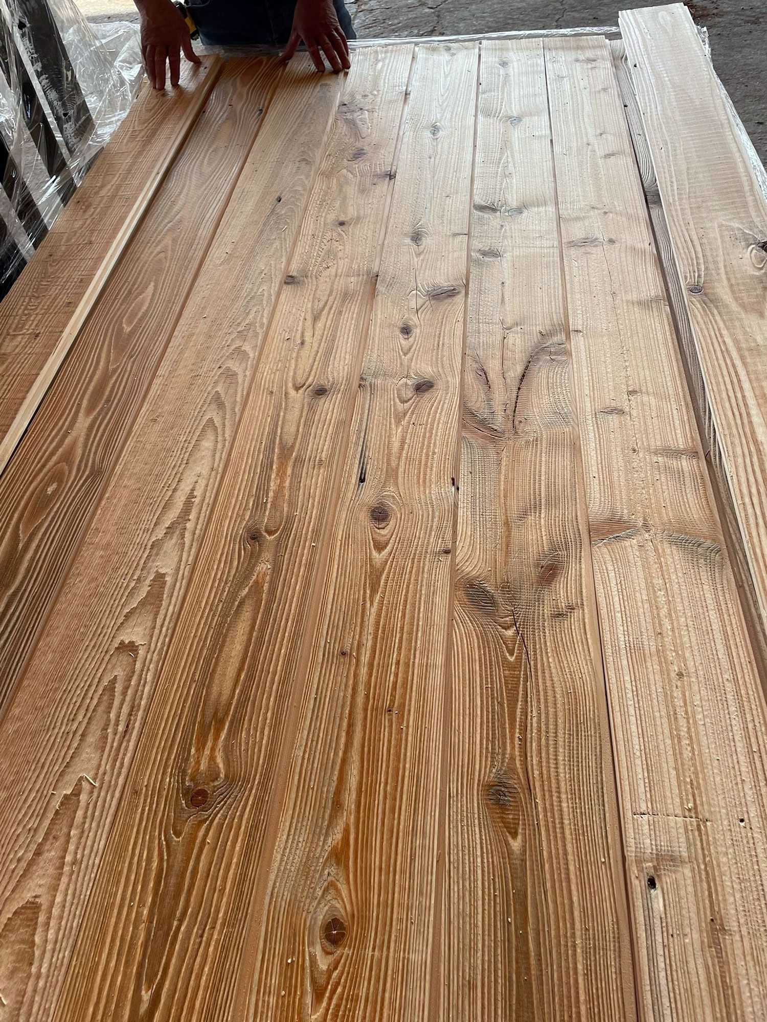 Tavole di legno disposte, alcune con nodi e venature visibili. In alto si vedono le mani di una persona.