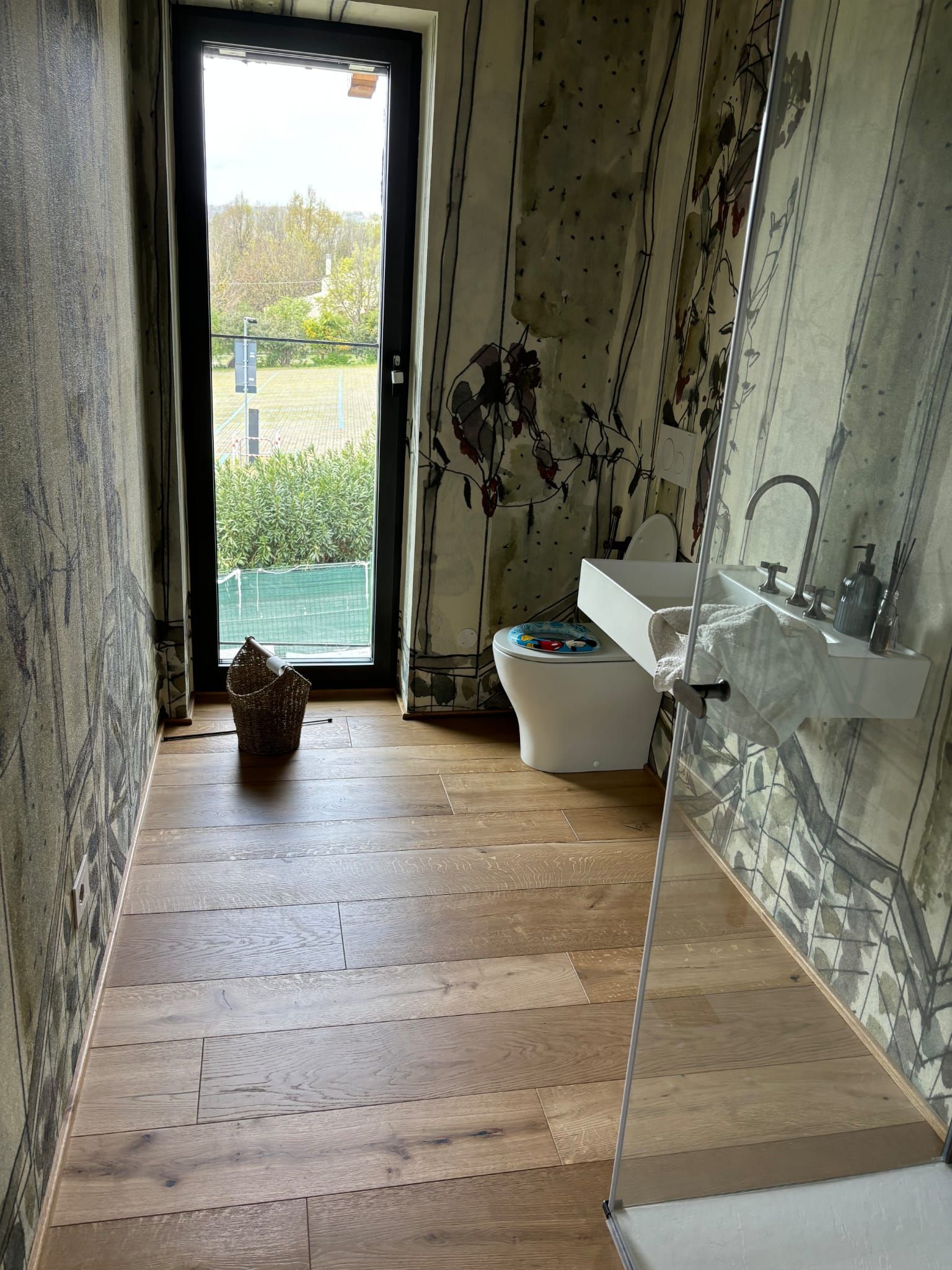 Bagno con pavimento in legno, pareti decorate, WC, lavandino, doccia in vetro e porta che dà sul cortile.