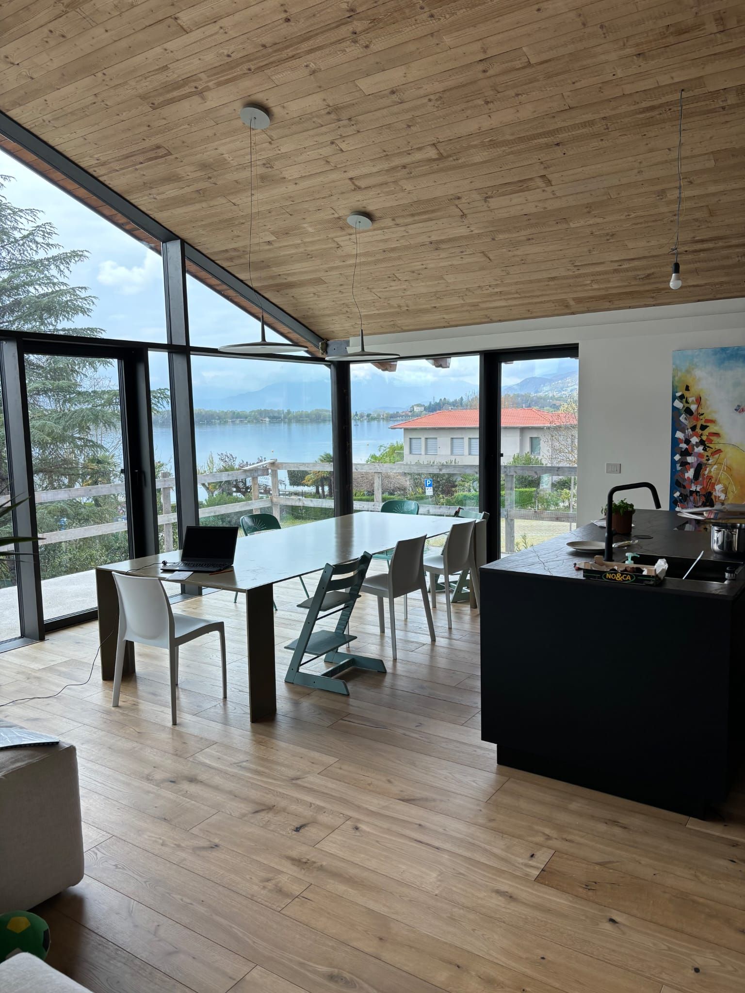 Sala da pranzo con ampie finestre che si affacciano sul lago. Tavolo con sedie, isola nera e soffitto in legno.