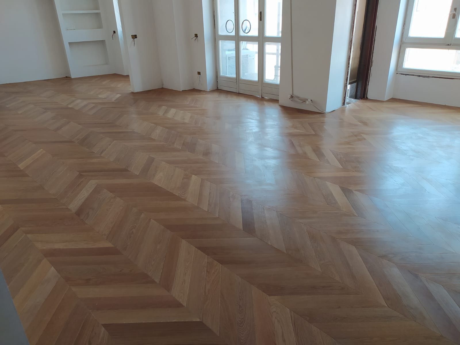Pavimento in parquet di legno chiaro in una stanza con pareti, finestre e porta bianche.