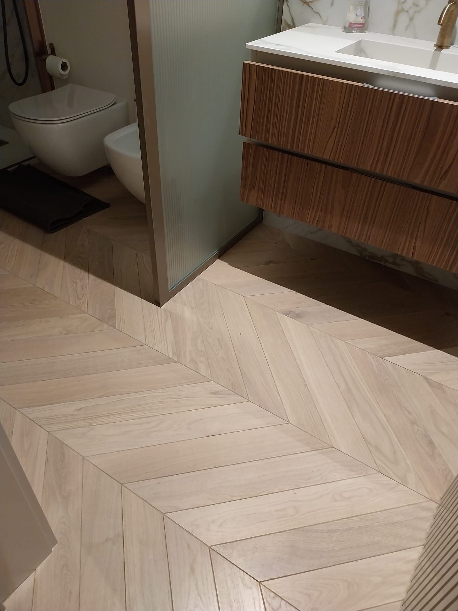 Pavimento in legno chiaro a spina di pesce in un bagno con WC e mobiletto in legno.