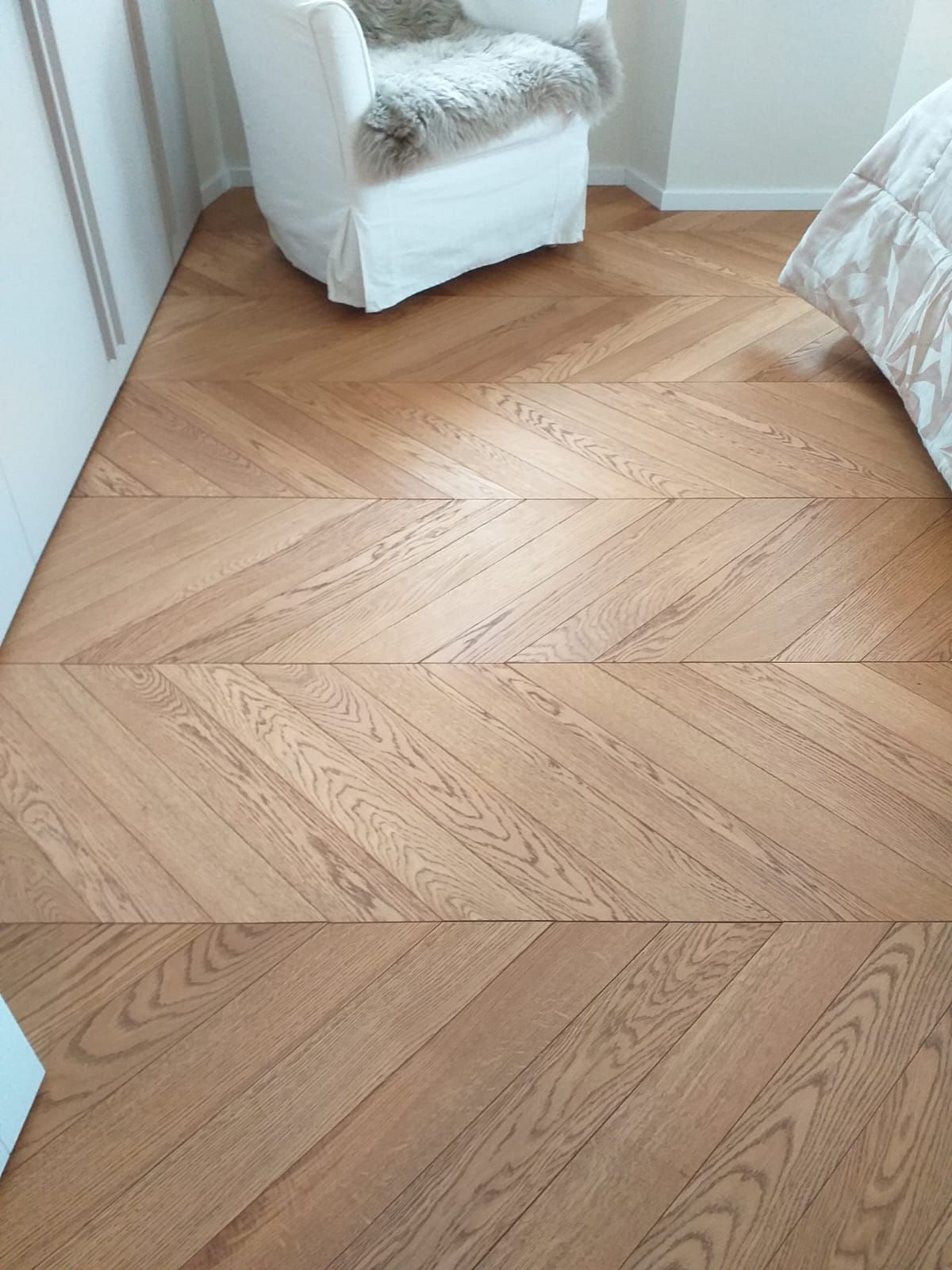 Pavimento in legno a spina di pesce in una stanza, con una sedia bianca e un letto parzialmente visibile.