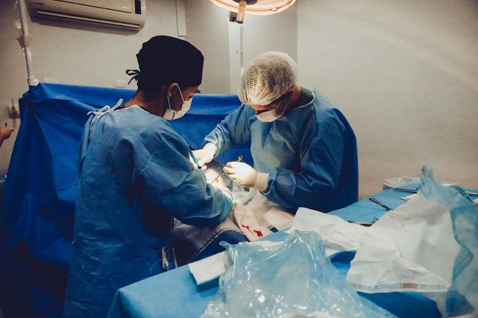 Two medical staff in blue gowns operate under bright lights on a patient in a clinical setting