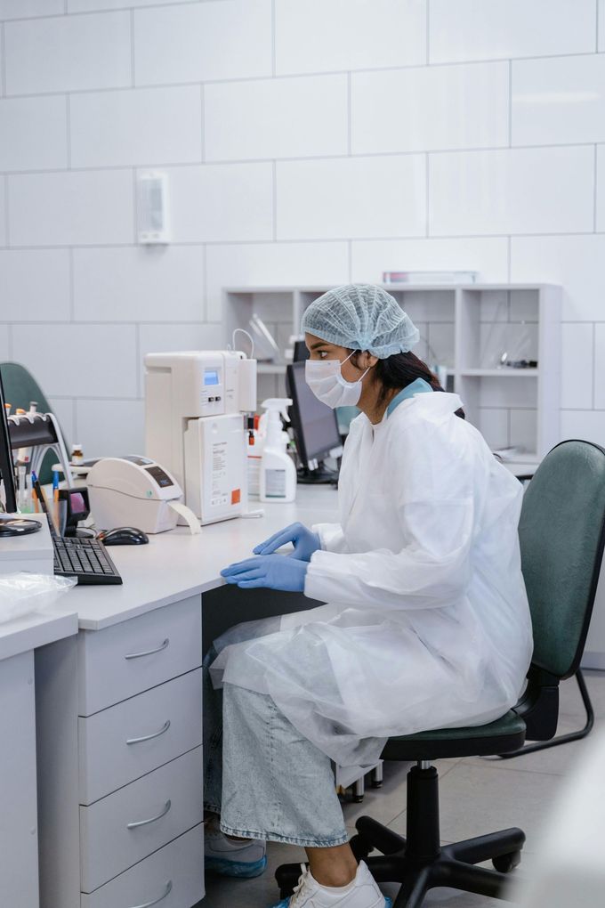 Lab worker in white coat and hairnet seated at a desk in a clinical workspace