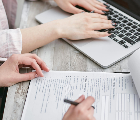 A person is typing on a laptop while another person fills out a form
