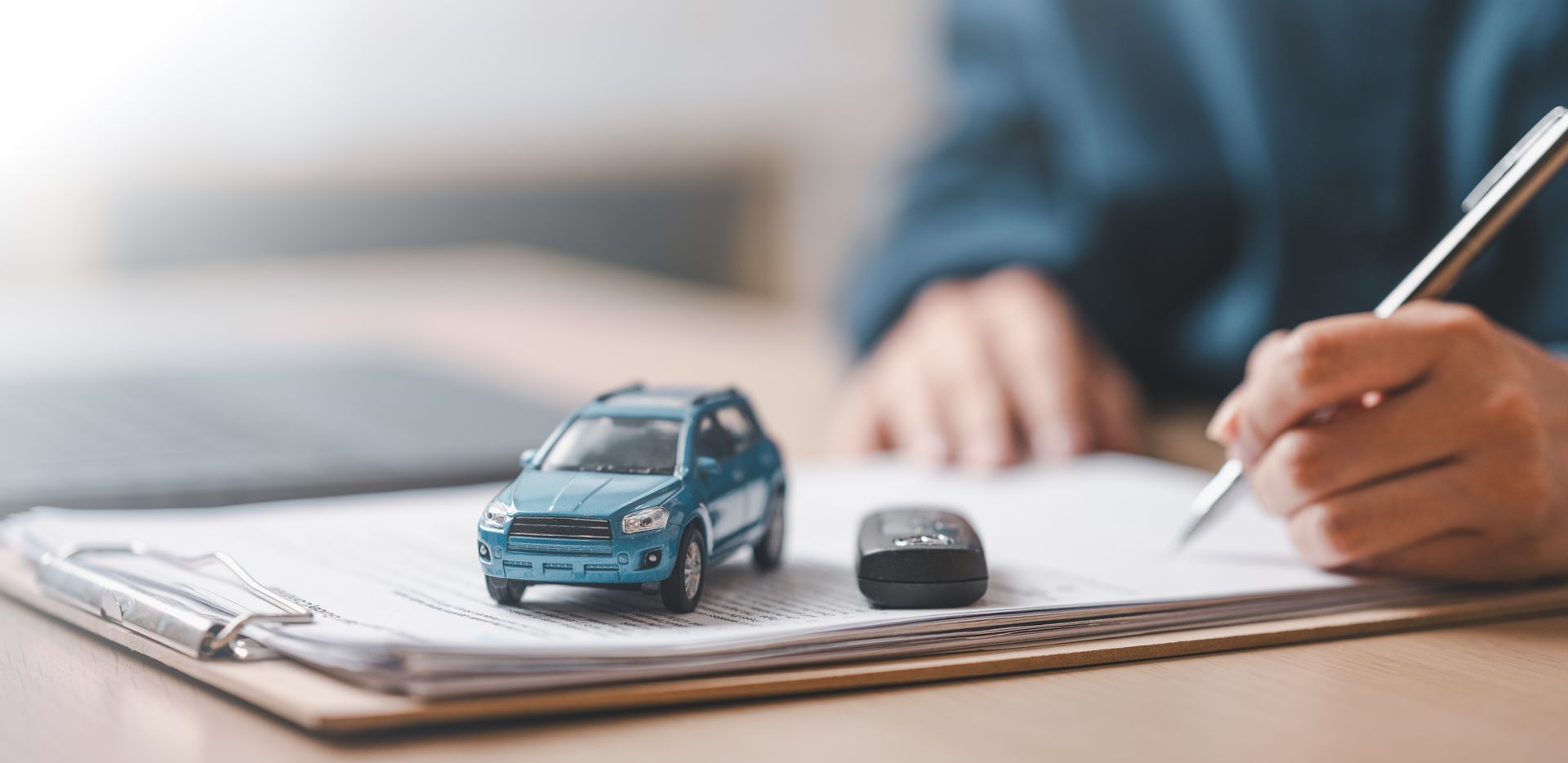 Miniature blue car and key fob on insurance documents as a person fills out paperwork.