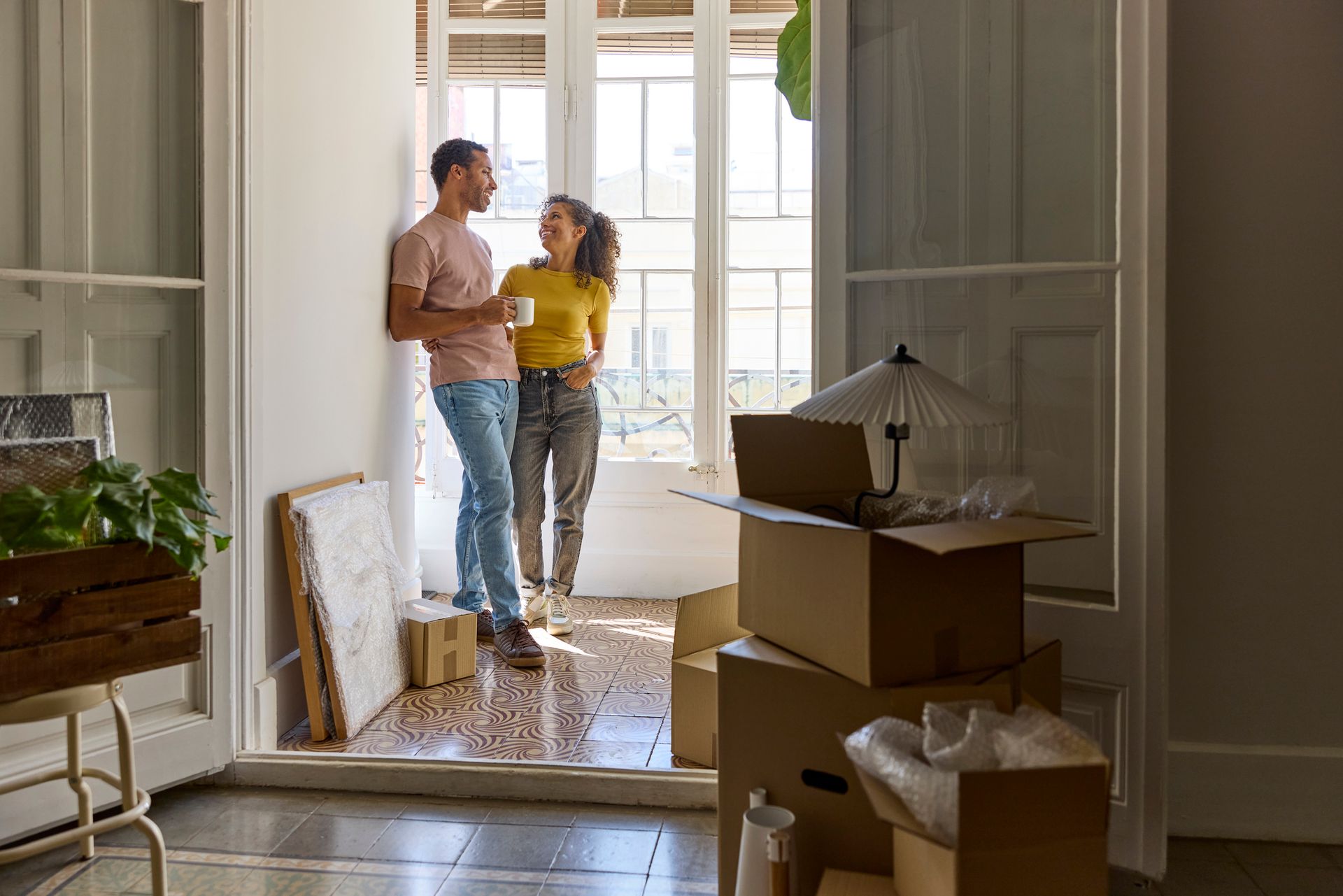 Couple stands in a doorway among moving boxes in a bright, partially unpacked room.